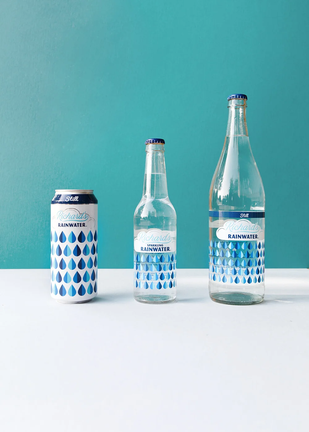 Three containers of Richard's Rainwater: a can, a small bottle, and a large bottle, all with blue raindrop patterns on the labels, set against a teal wall background.