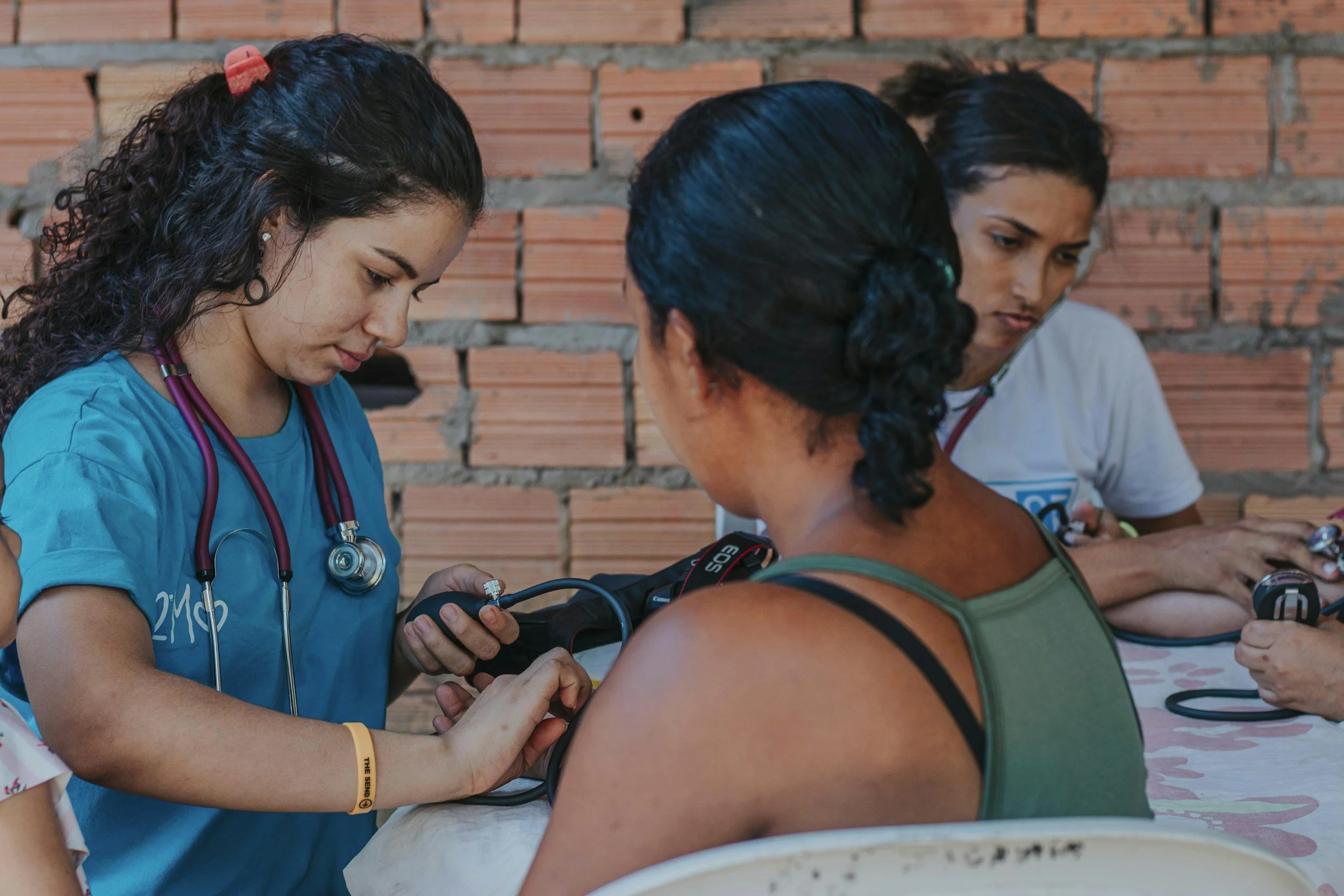 Healthcare workers in scrubs and scrubs and stethoscopes are examining a woman with a blood pressure cuff in an outdoor setting with a brick wall in the background.