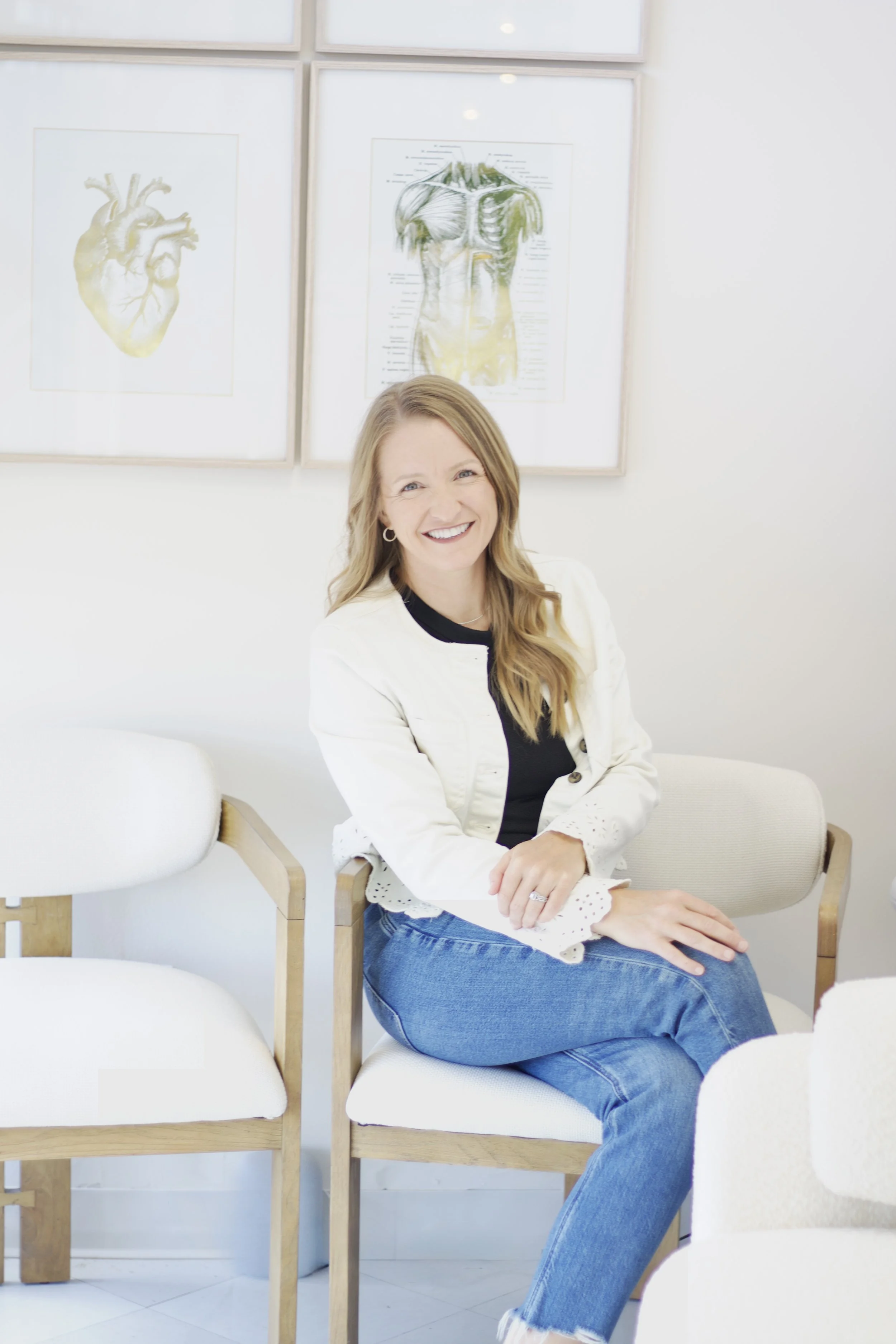 A woman with long blonde hair smiling and sitting on a white chair in a white room with framed anatomical posters of the human heart and muscular system on the wall behind her.