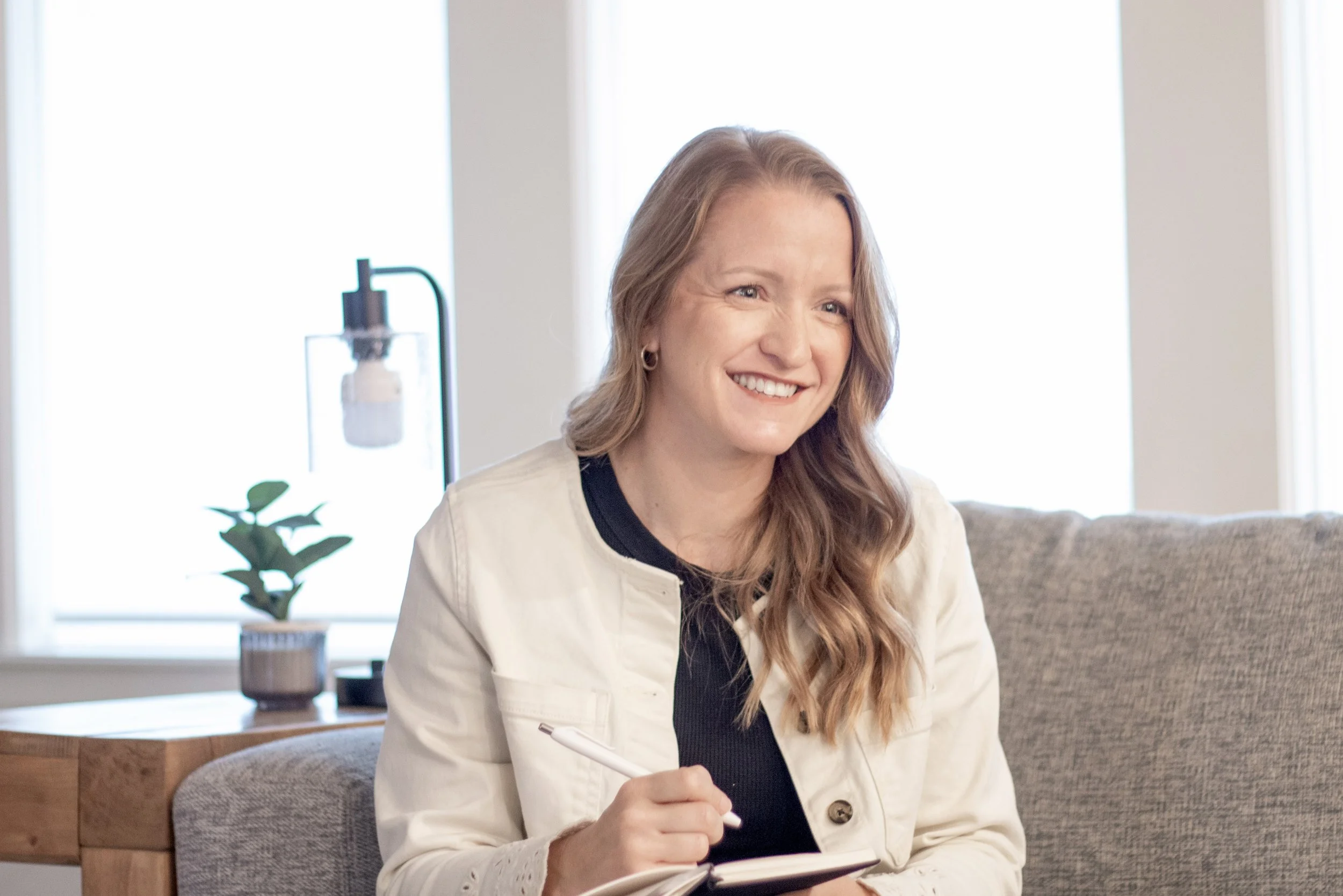 A woman with long, wavy, reddish hair, smiling warmly, is sitting on a beige sofa. She is wearing a white blazer over a black top and holding a pen and notebook, possibly in a cozy, well-lit indoor space.