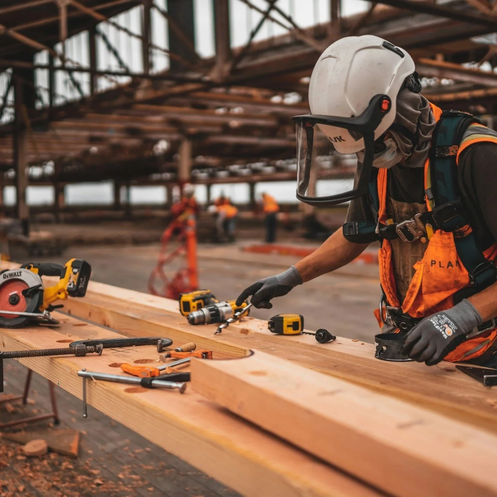 A construction worker wearing protective gear, including a white helmet with a face shield and an orange safety vest, working on a wooden structure with various tools and equipment on a workbench in a construction site.