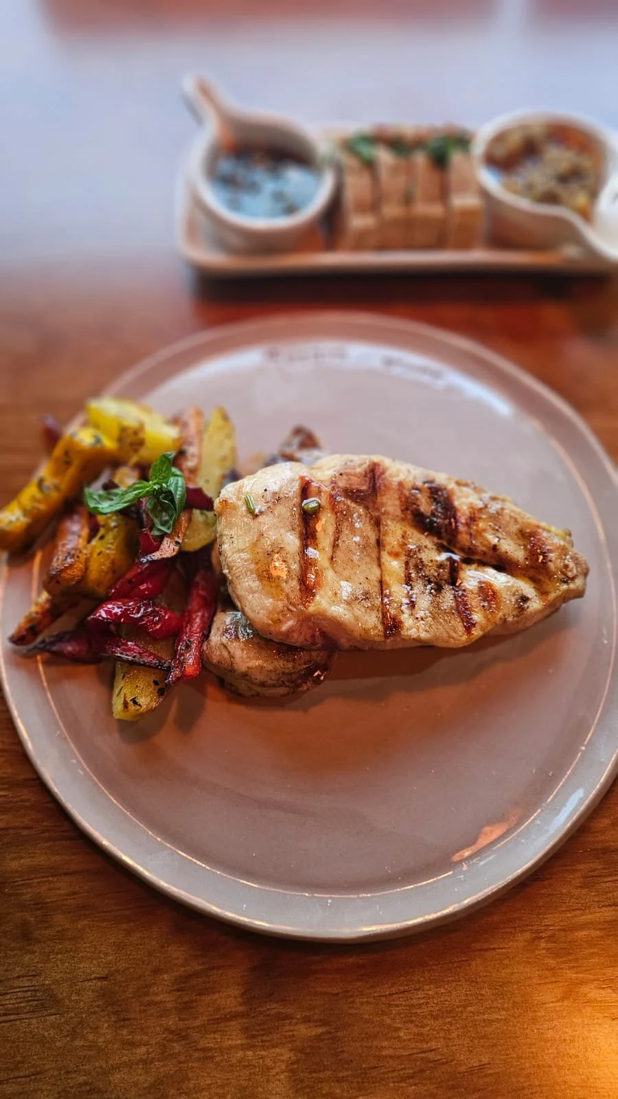 Grilled chicken breast with roasted vegetables on a plate, with dipping sauces in the background.