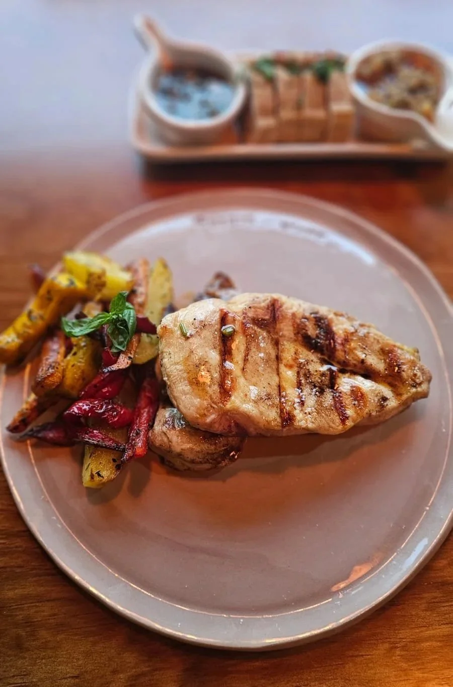 Grilled chicken breast with roasted vegetables on a plate and small bowls of sauces or dips in the background.