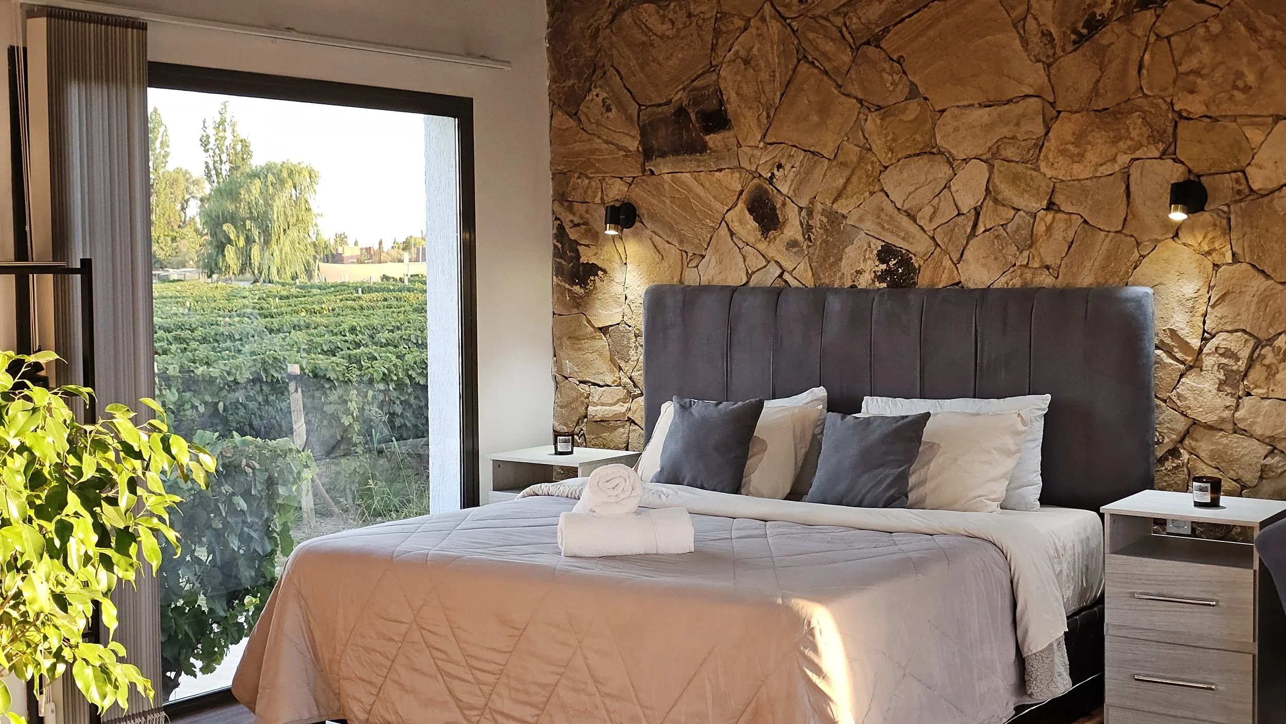 A bedroom with a stone accent wall, a large bed with a gray headboard, and white and gray pillows. There are two bedside tables, each with a small candle. A large window shows a view of greenery and trees outside, and sunlight streams into the room. A green plant is partially visible on the left side.