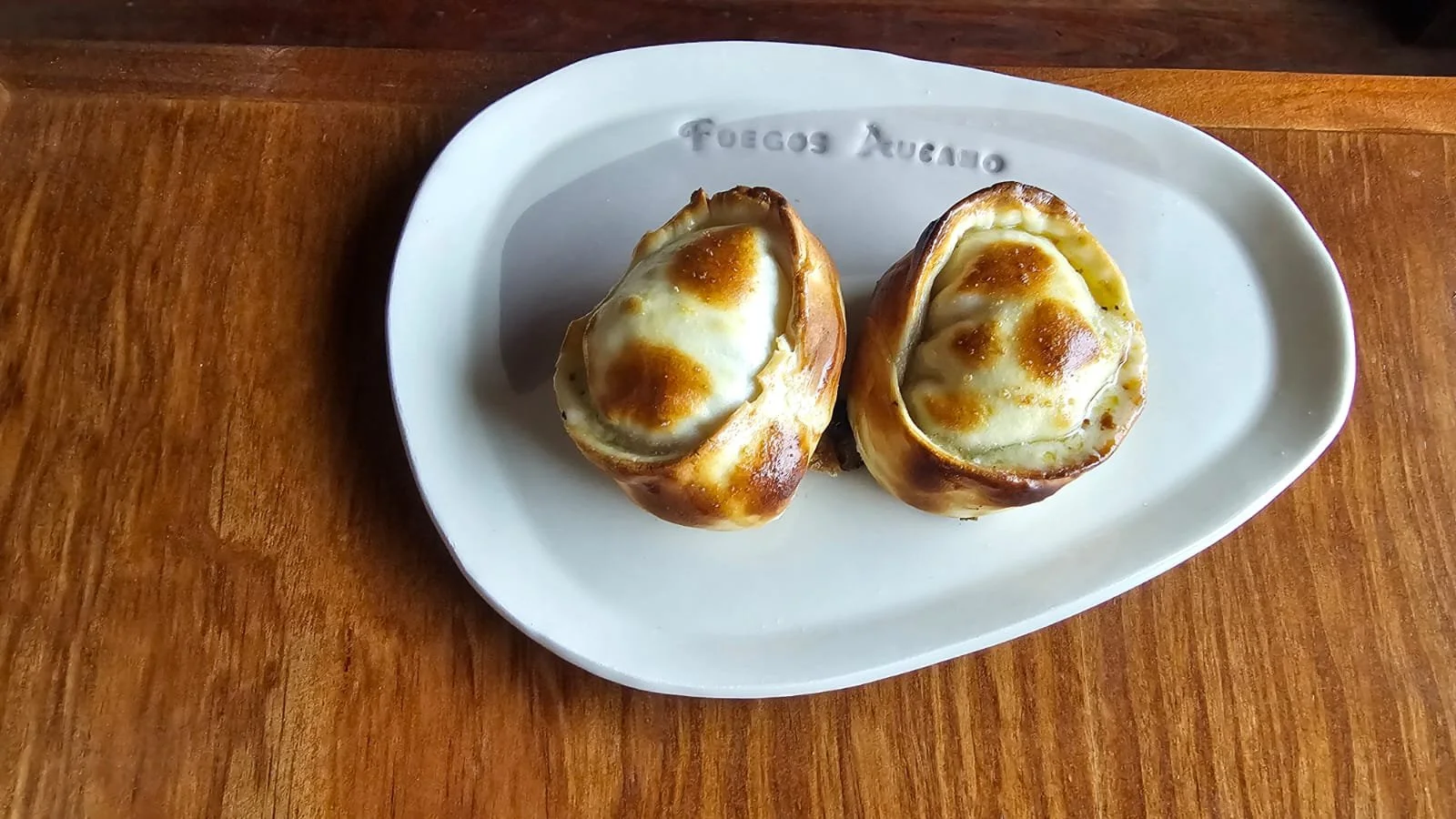 Two stuffed baked eggs with melted cheese on a white oval plate with engraved text, placed on a wooden surface.