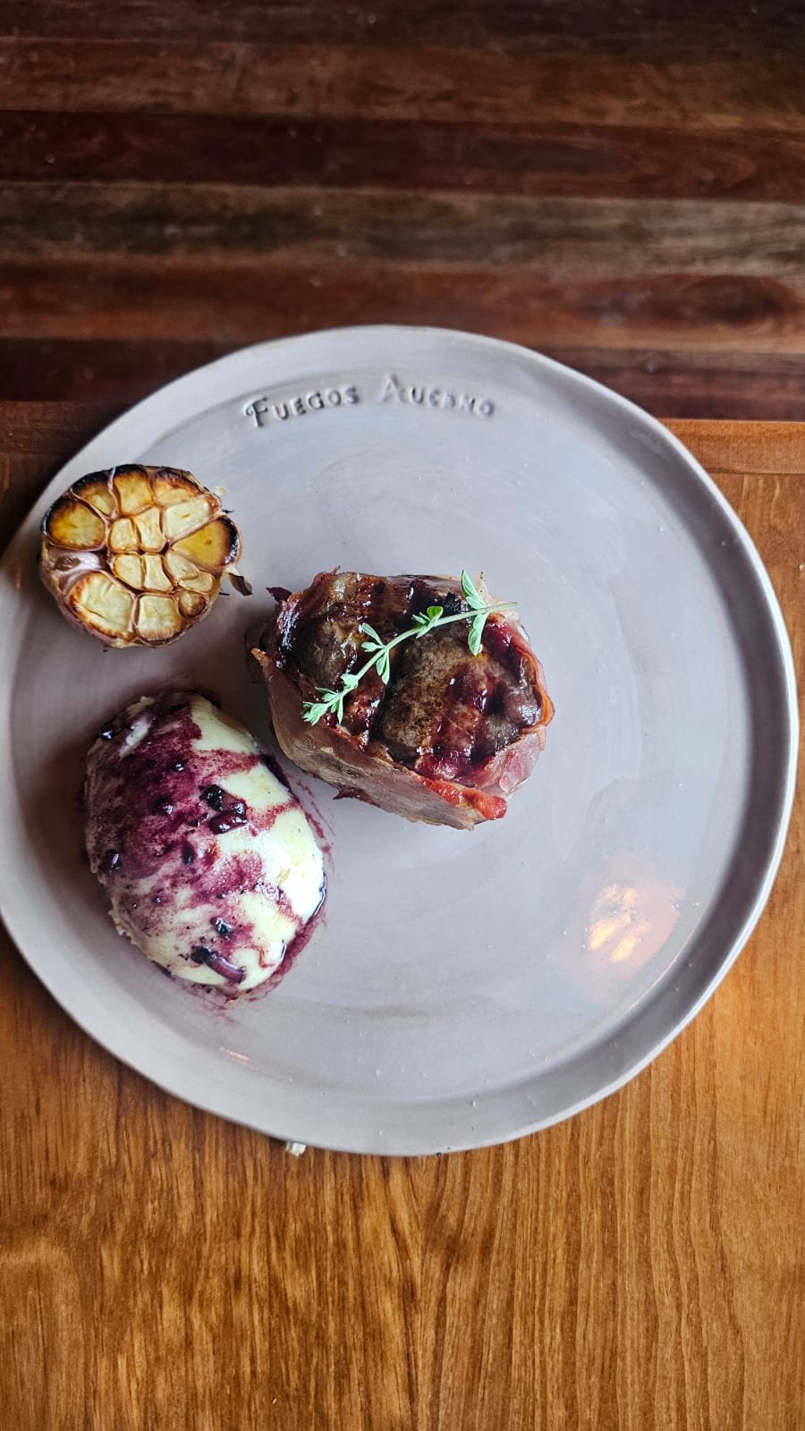 A ceramic plate with the words "Fuegos Argentinos" contains a serving of meat, a roasted garlic bulb, and a scoop of ice cream with berry sauce, garnished with a small sprig of herbs.
