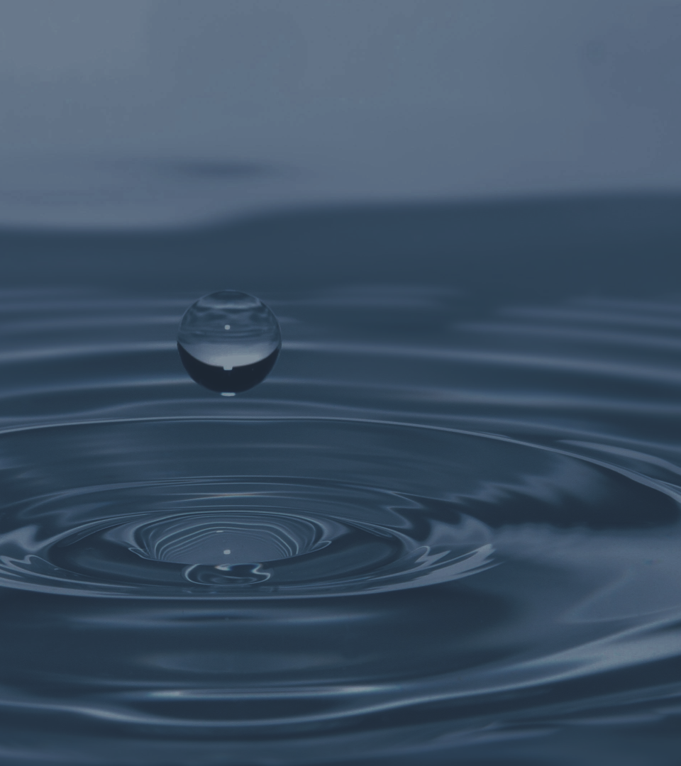 A close-up of a water surface with a single water droplet mid-air just above the ripples.