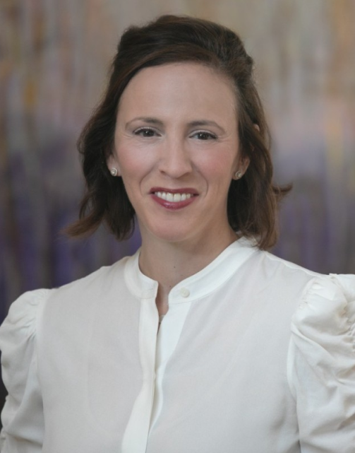 A woman with shoulder-length brown hair, wearing a white top, smiling at the camera with a blurred outdoor background.
