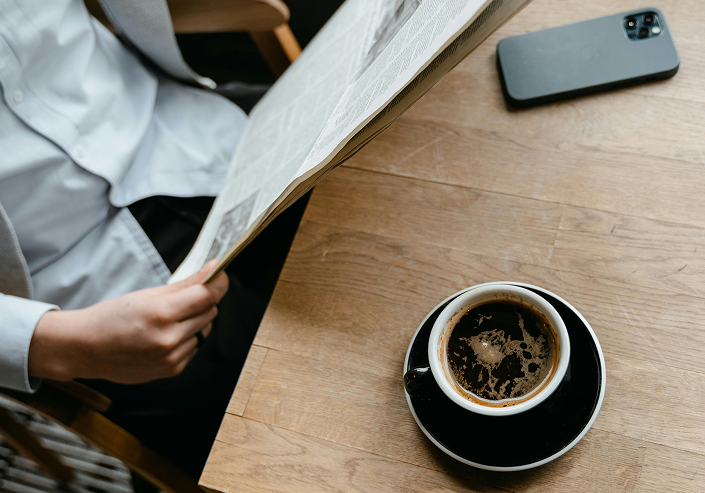 Person reading a newspaper at a wooden table with a cup of black coffee and a smartphone.