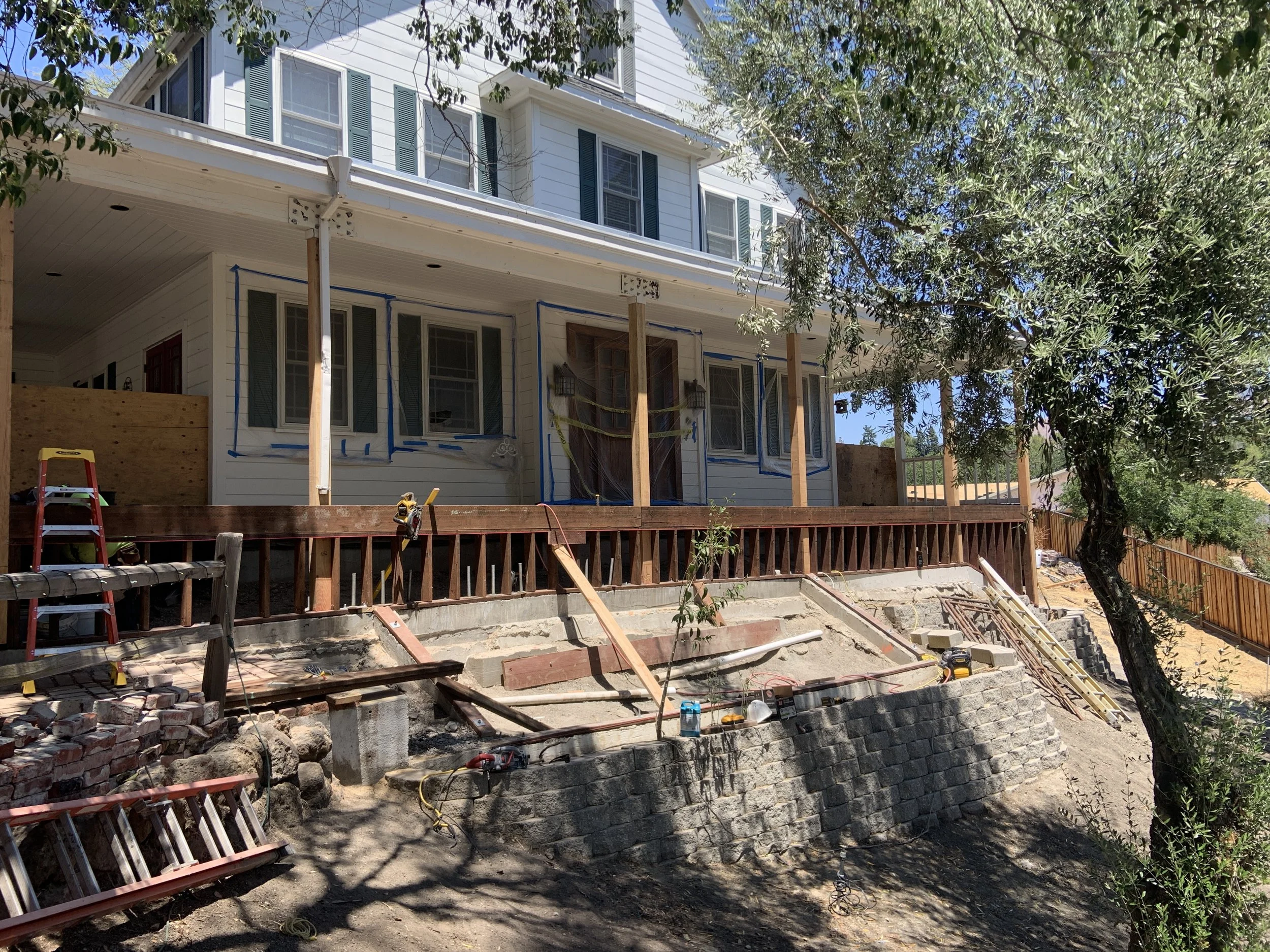 A house under renovation with a wooden deck becoming a stairway, construction tools and materials, and a tree in the foreground.