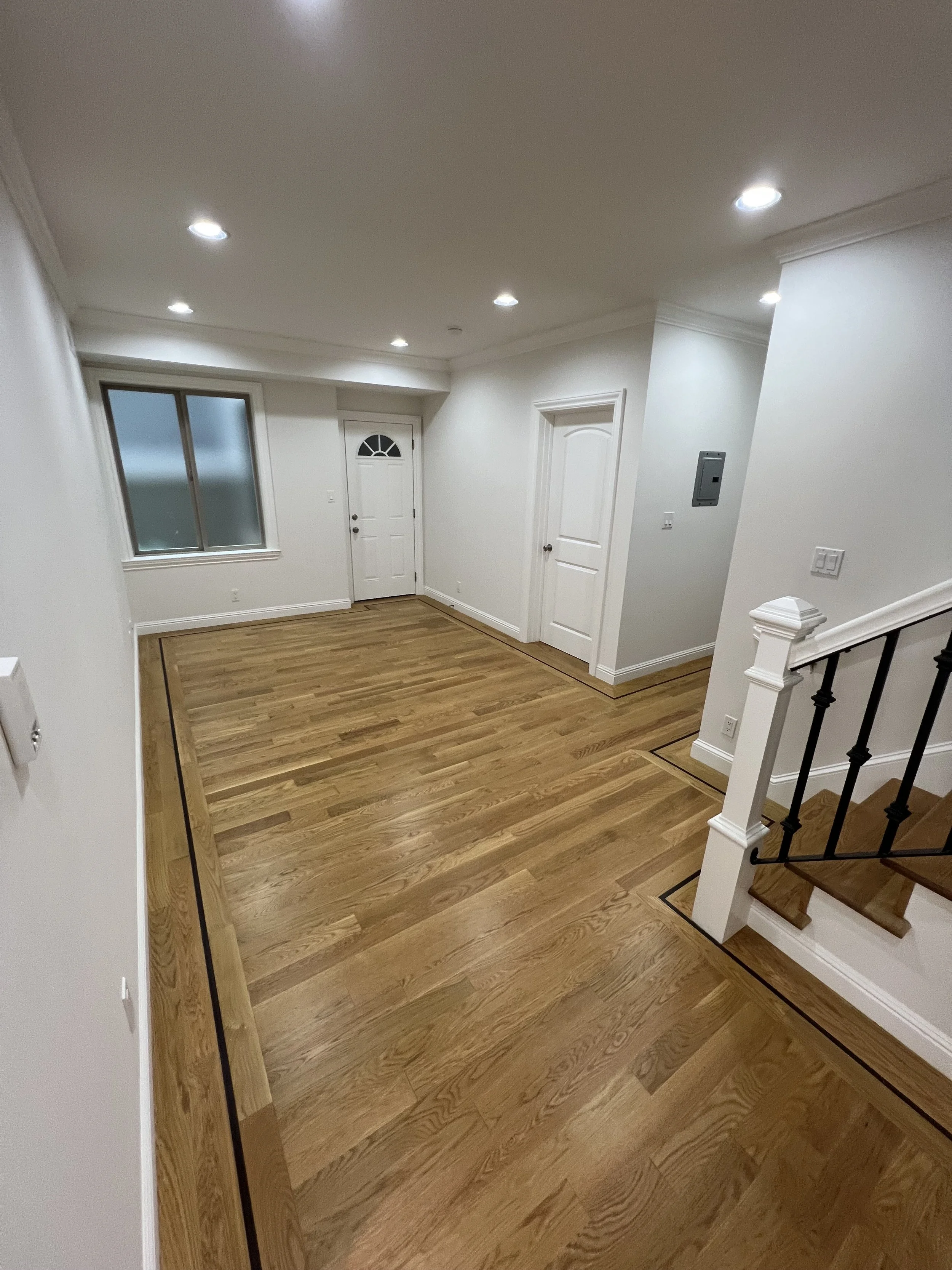 Empty living room with wooden flooring, white walls, a window, and a white front door. Recessed lighting and staircase with black railing visible.