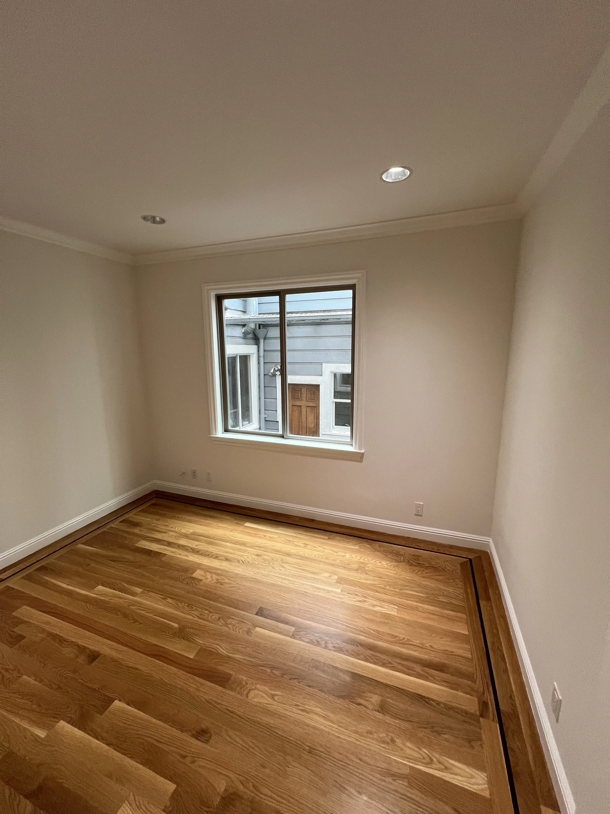 Empty room with hardwood floor, beige walls, white trim, and a single window showing neighboring buildings.