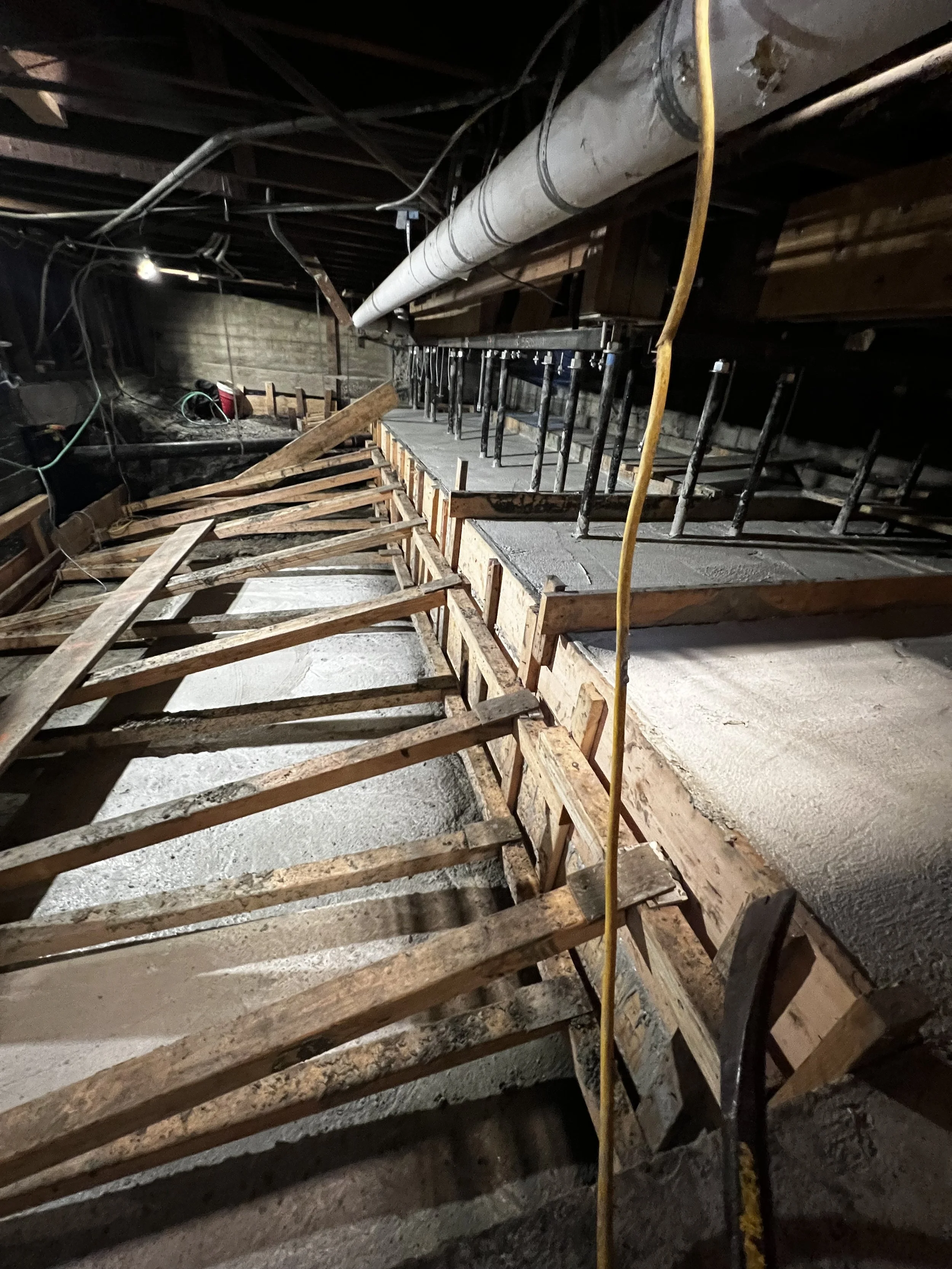 Construction site with wooden formwork and concrete being poured for a staircase, with pipes and electrical wiring overhead.