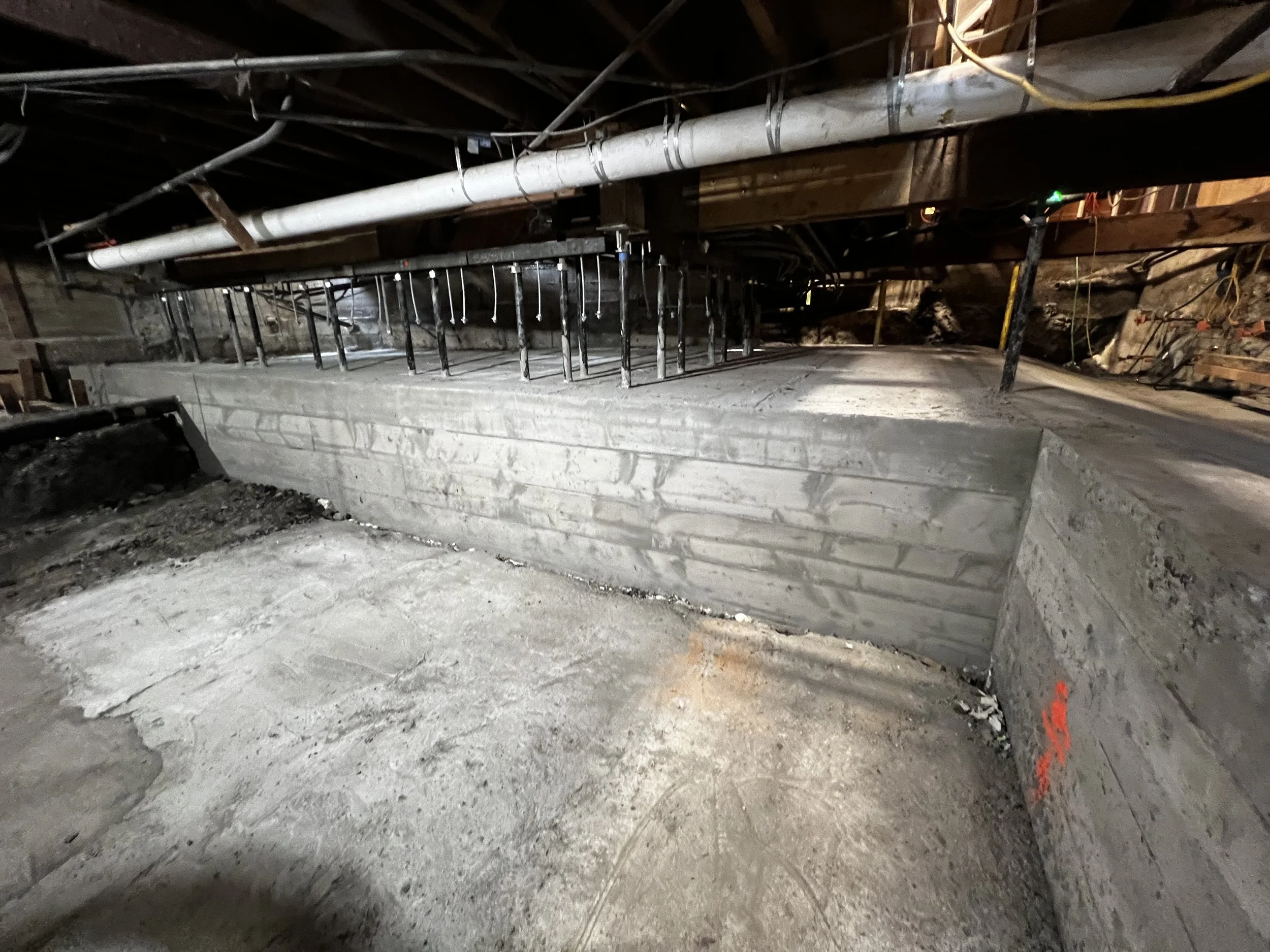 Construction site with freshly poured concrete foundation and rebar for reinforcement, visible pipes and support beams overhead.