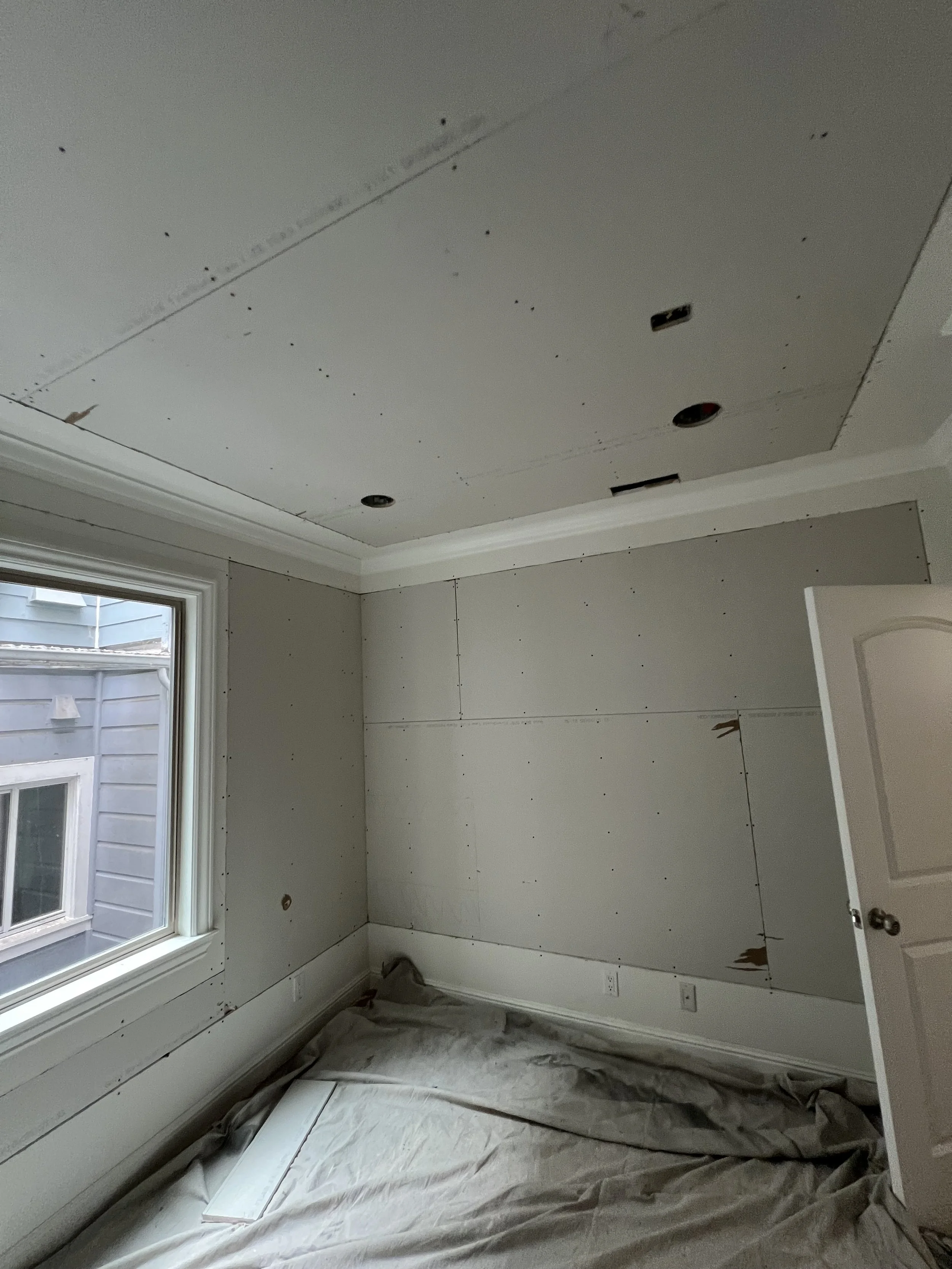 Empty room under construction with drywall in the process of being installed, a window on the left, a partially open door on the right, and a beige tarp covering the floor.