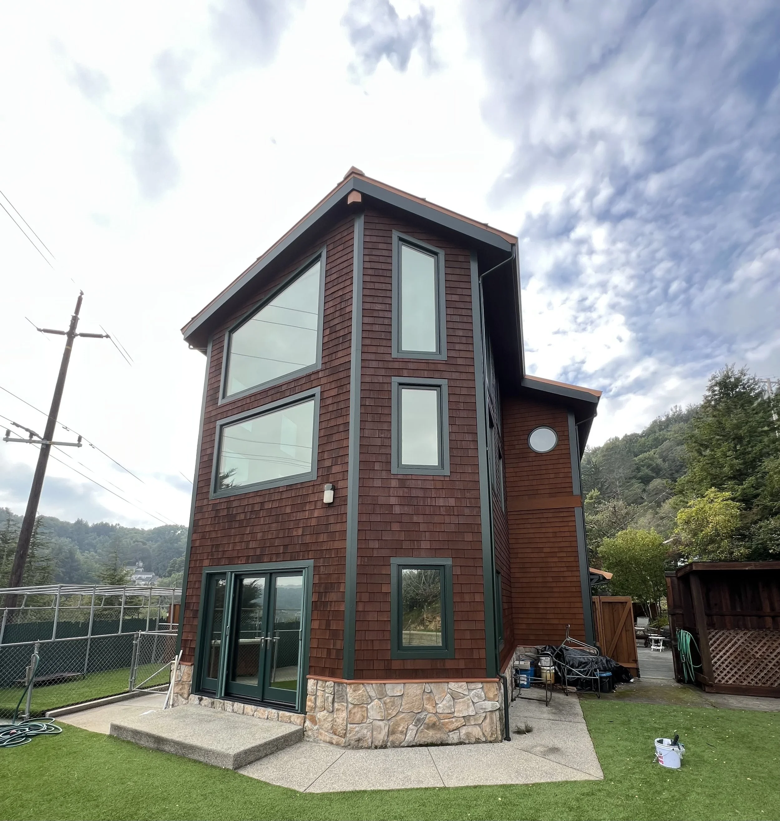 A modern multi-story house with wooden siding, multiple large windows, a stone foundation, a glass door, and a small outdoor patio area, situated in a green landscape with trees and mountains in the background.
