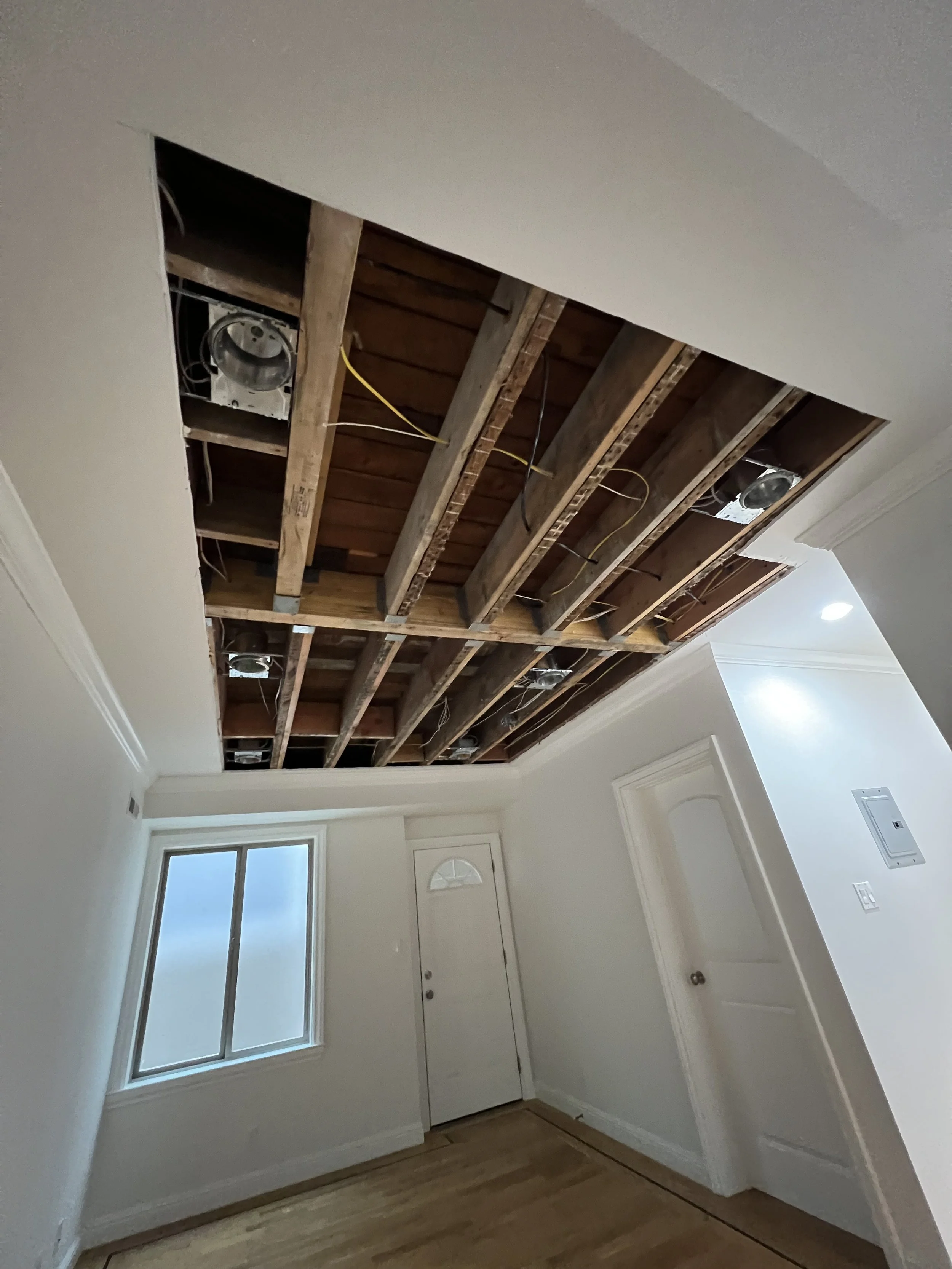 Living room with ceiling being renovated, showing exposed wooden beams and wiring, with a window on the left and a white door on the back wall.
