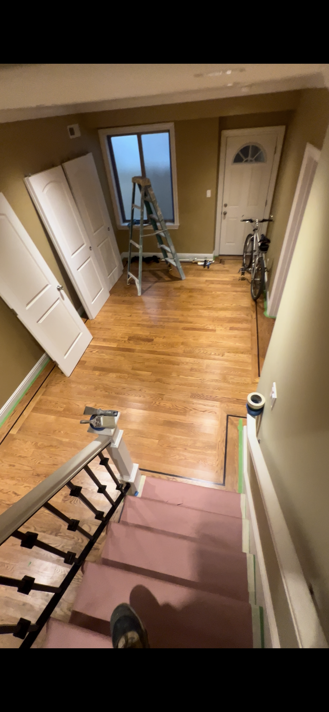 View from the top of a staircase looking into an indoor room with hardwood floors. The room has a window, a front door, a bicycle, a ladder, and open closet doors. The staircase has pink protective cover on the steps, and green painter's tape is along the baseboards and edges of the stairs.
