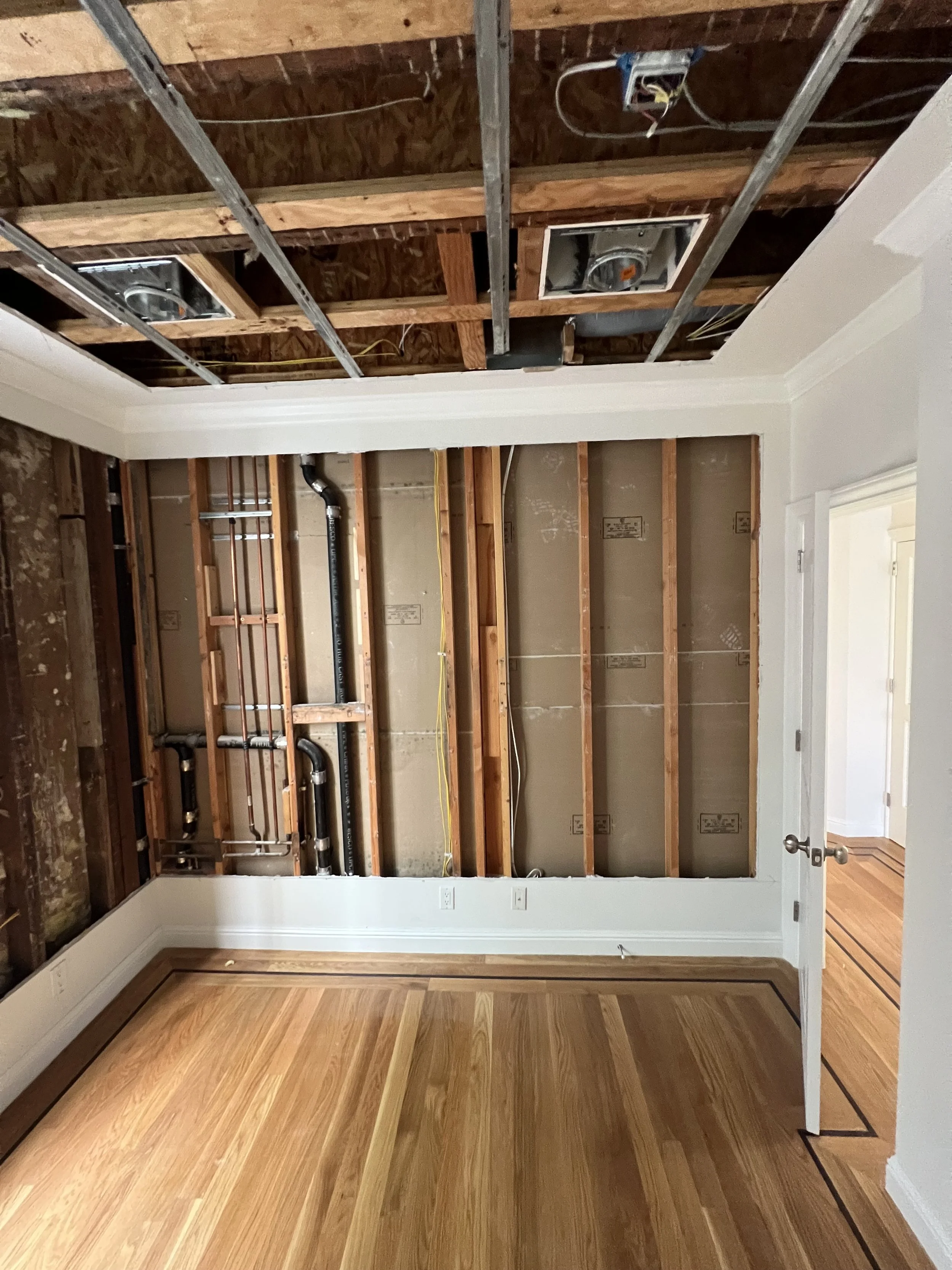 Room under renovation showing exposed ceiling with metal framing, pipes, and electrical wiring, with a finished hardwood floor and white trim.