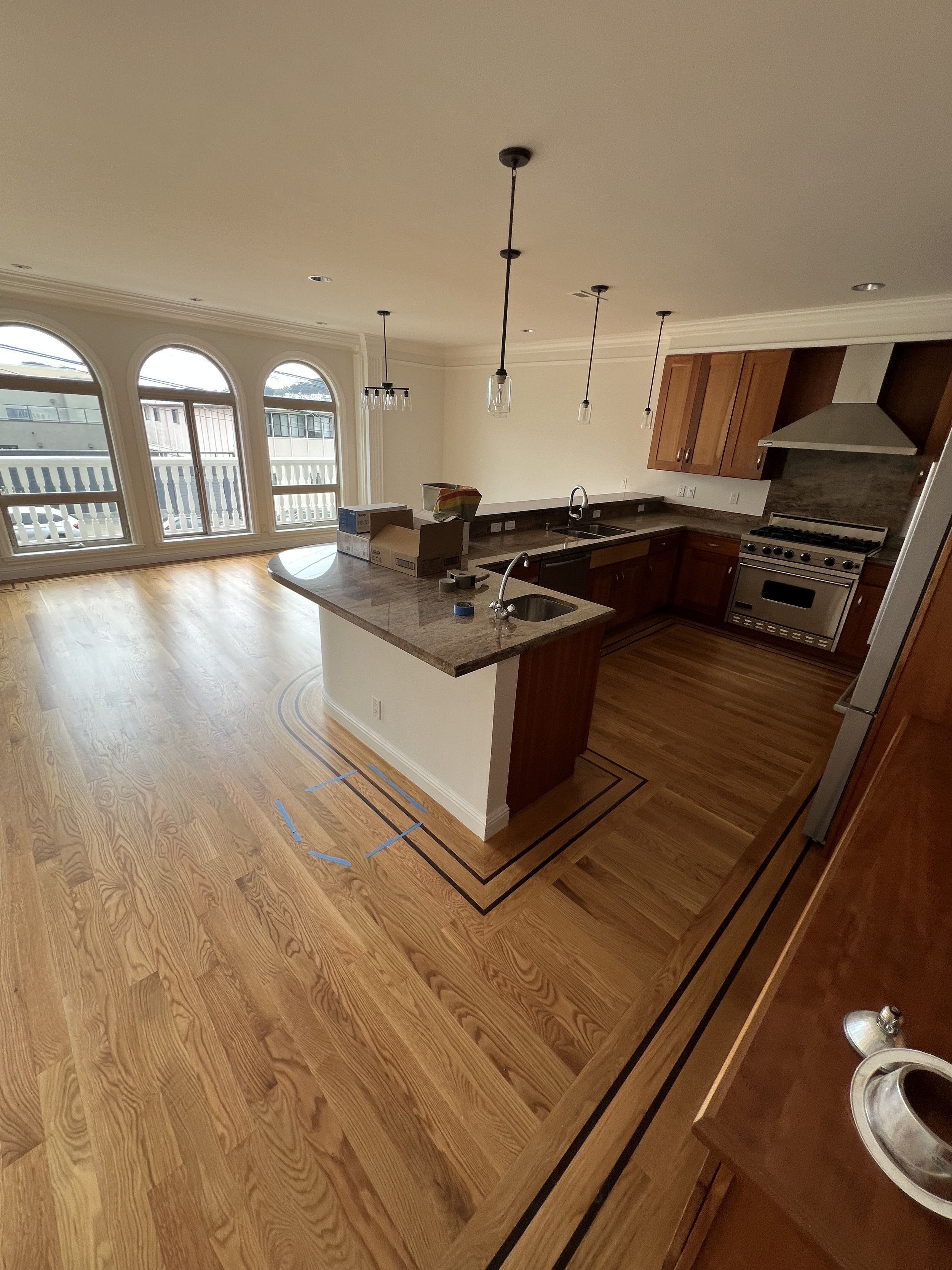 Empty kitchen with wooden flooring, a marble island, stainless steel stove, and large arched windows.