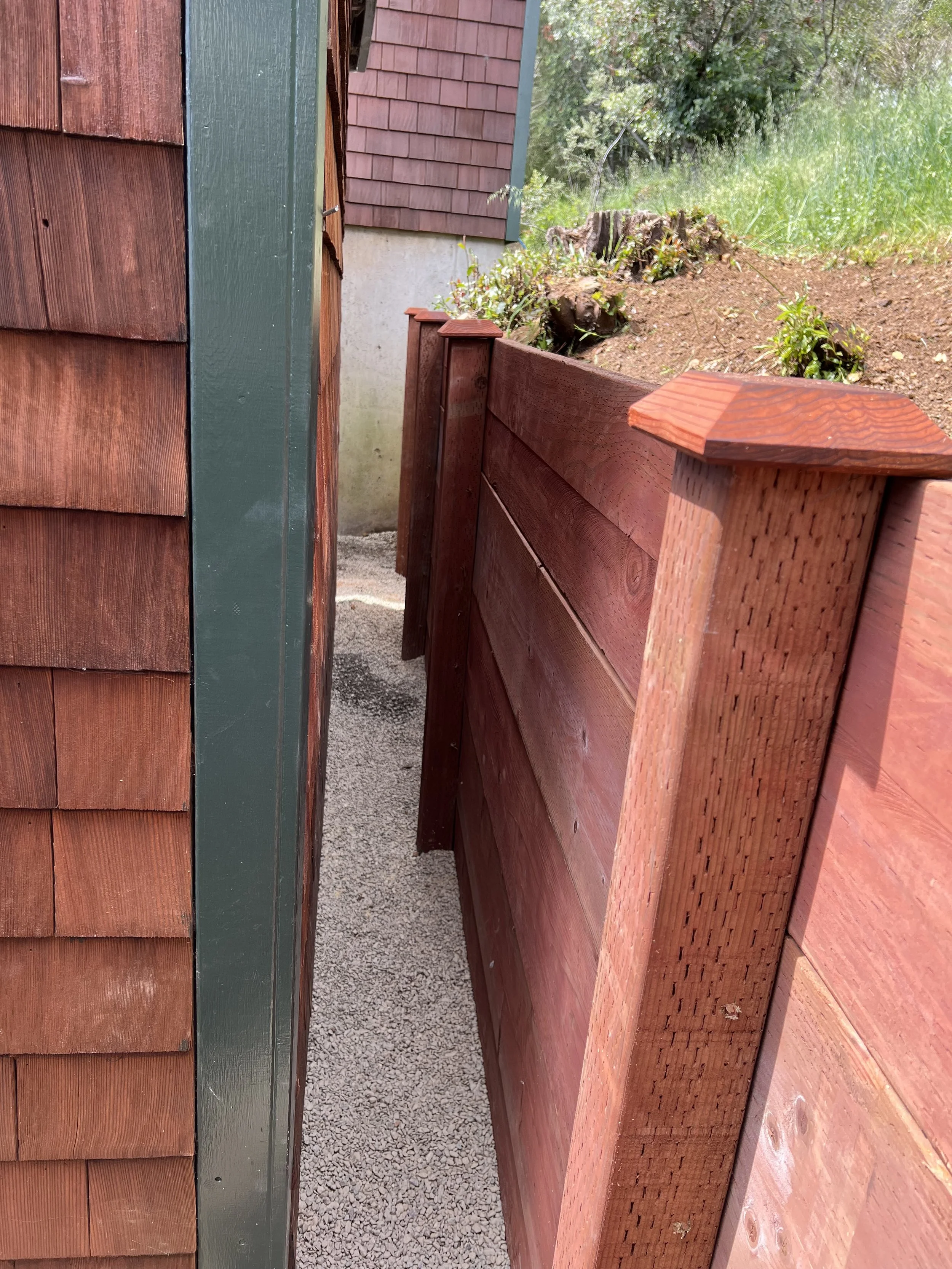 A narrow gravel pathway between two wooden fences attached to a house with brown siding, with green outdoors in the background.