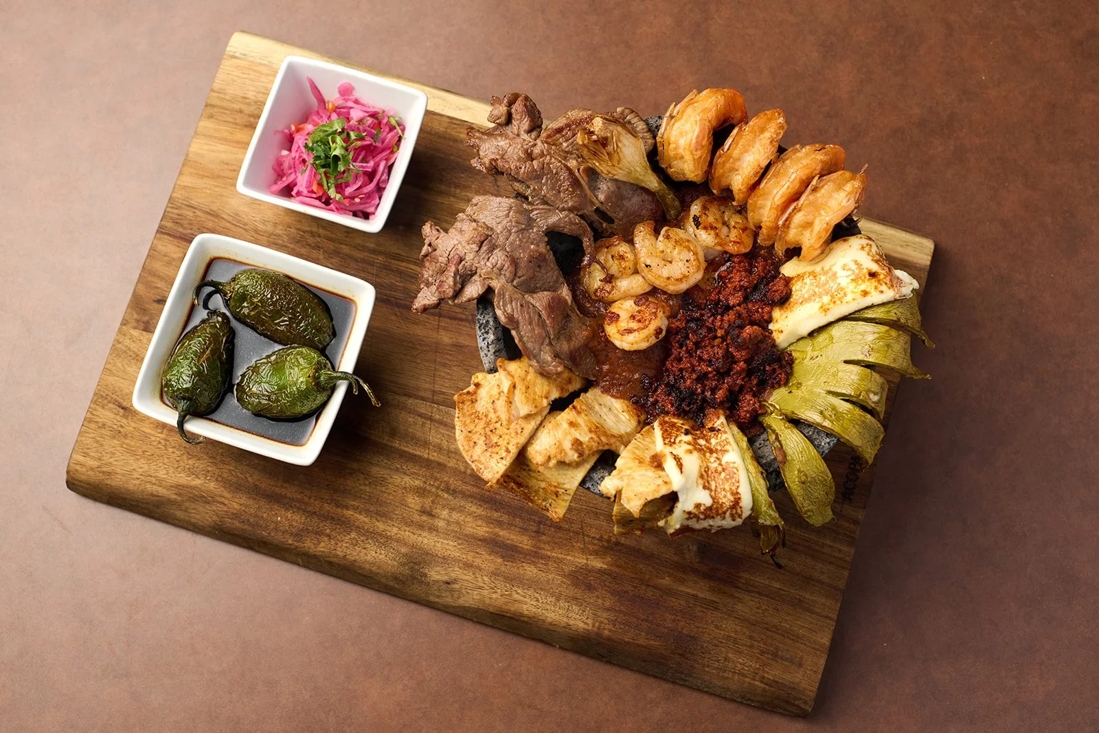 A wooden serving board with a variety of grilled meats and vegetables, including grilled cherry tomatoes, peppers, and chunks of chicken and beef, accompanied by two small dishes with pickled red cabbage and stuffed jalapeño peppers.