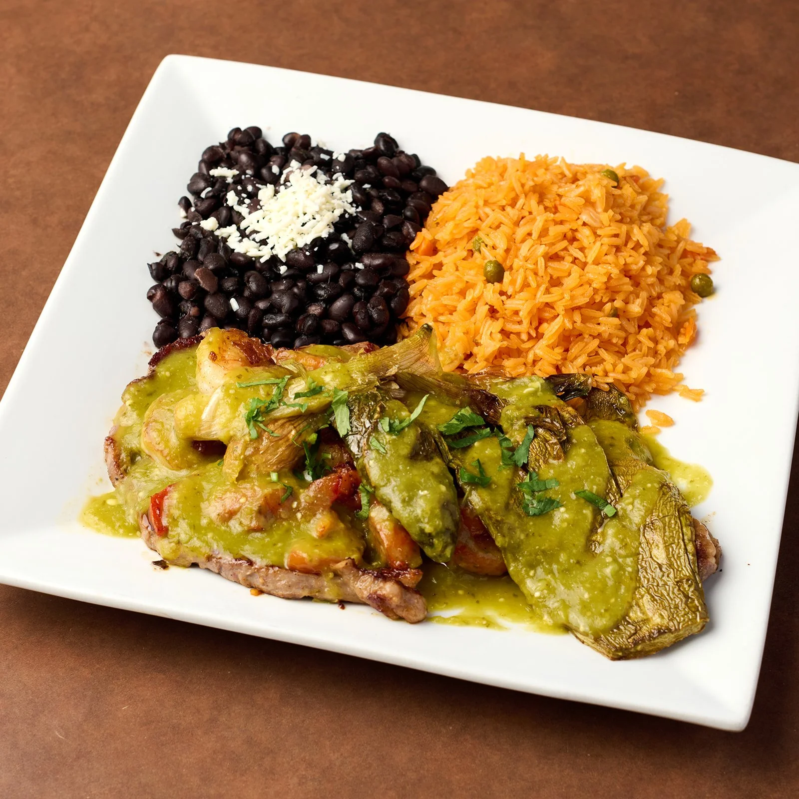 Plate of Mexican food with black beans topped with cheese, rice, and a cooked meat dish with green sauce and vegetables.