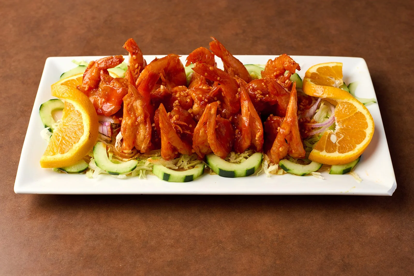 Rectangular plate of fried shrimp with orange slices and sliced cucumbers on a bed of shredded cabbage and lettuce