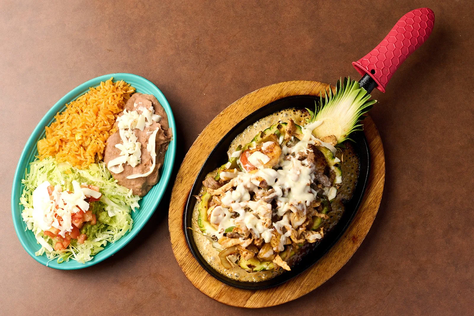 A sizzling dish of grilled meat topped with melted cheese, served on a pineapple leaf with sliced vegetables. Next to it, a bowl of rice, refried beans with cheese, and shredded lettuce with tomatoes on a turquoise plate.