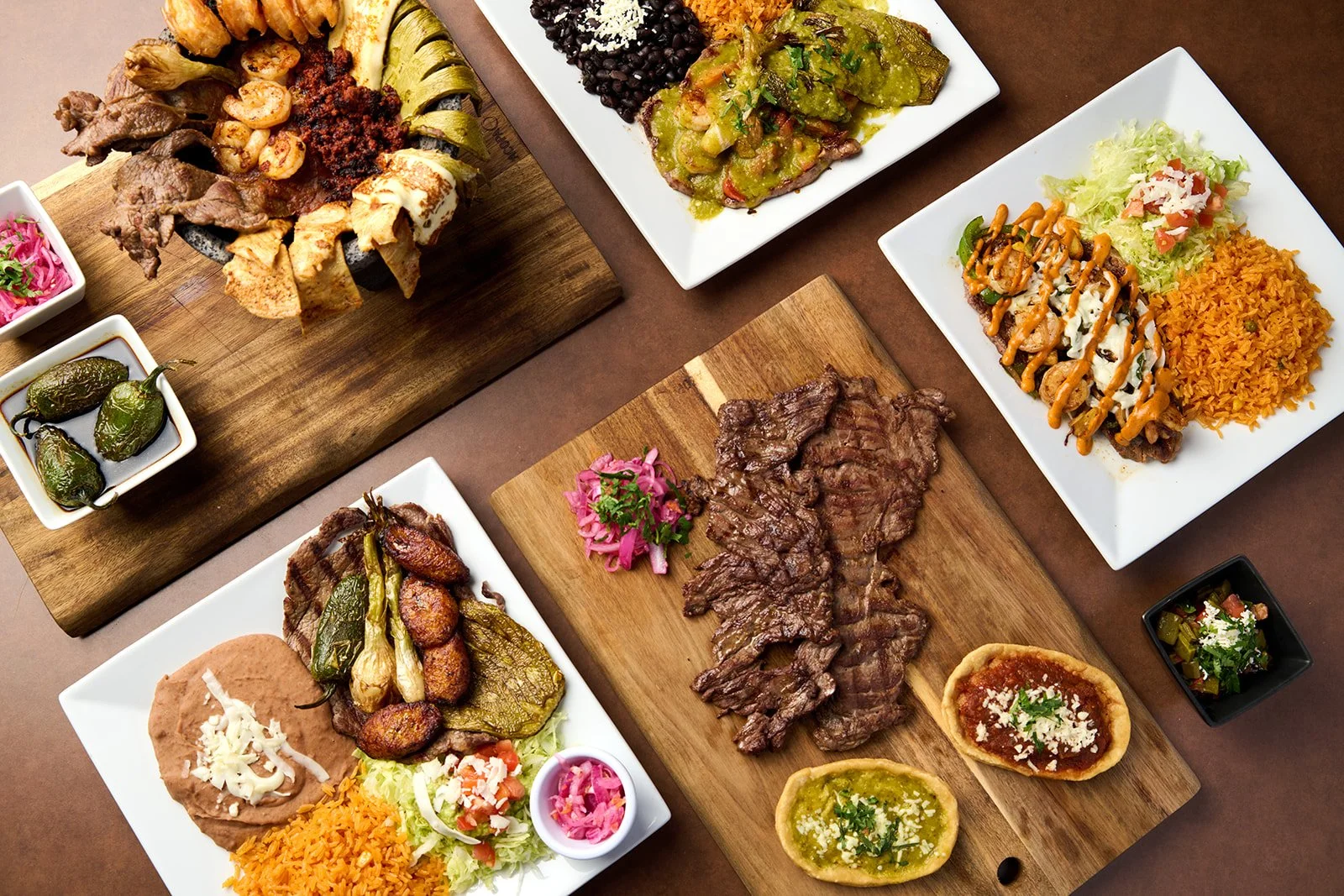 Assorted Mexican dishes on wooden and white plates, including grilled beef, chicken, and steak with vegetables, rice, beans, and traditional sides like pickled onions, peppers, and salsas.