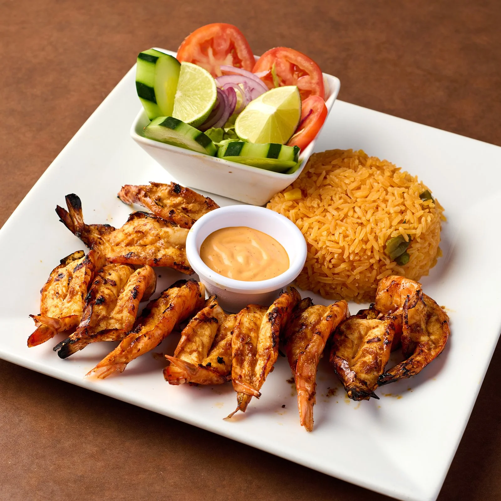 Plate of grilled shrimp with orange rice, side salad with tomato, cucumber, lemon, and onion, and dipping sauce.