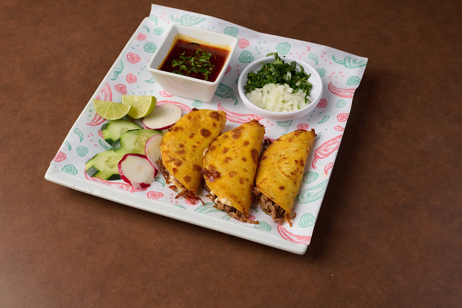 Plate with three beef-filled tacos, lime wedges, sliced radishes, cucumbers, chopped onions, chopped cilantro, and small bowls of red salsa and chopped onions with herbs.