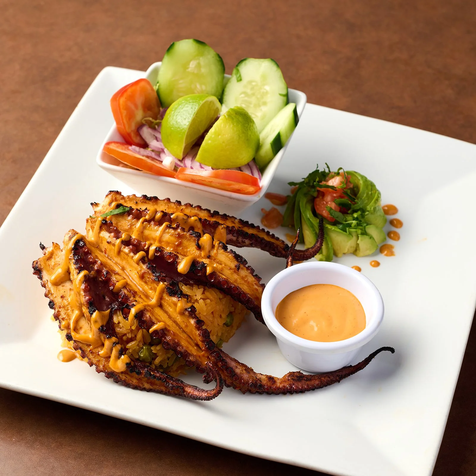 Grilled octopus with seasoned rice, topped with sauce, served with side salad of cucumbers, tomatoes, red onions, avocado, and lime slices, accompanied by a small bowl of dipping sauce on a white plate.