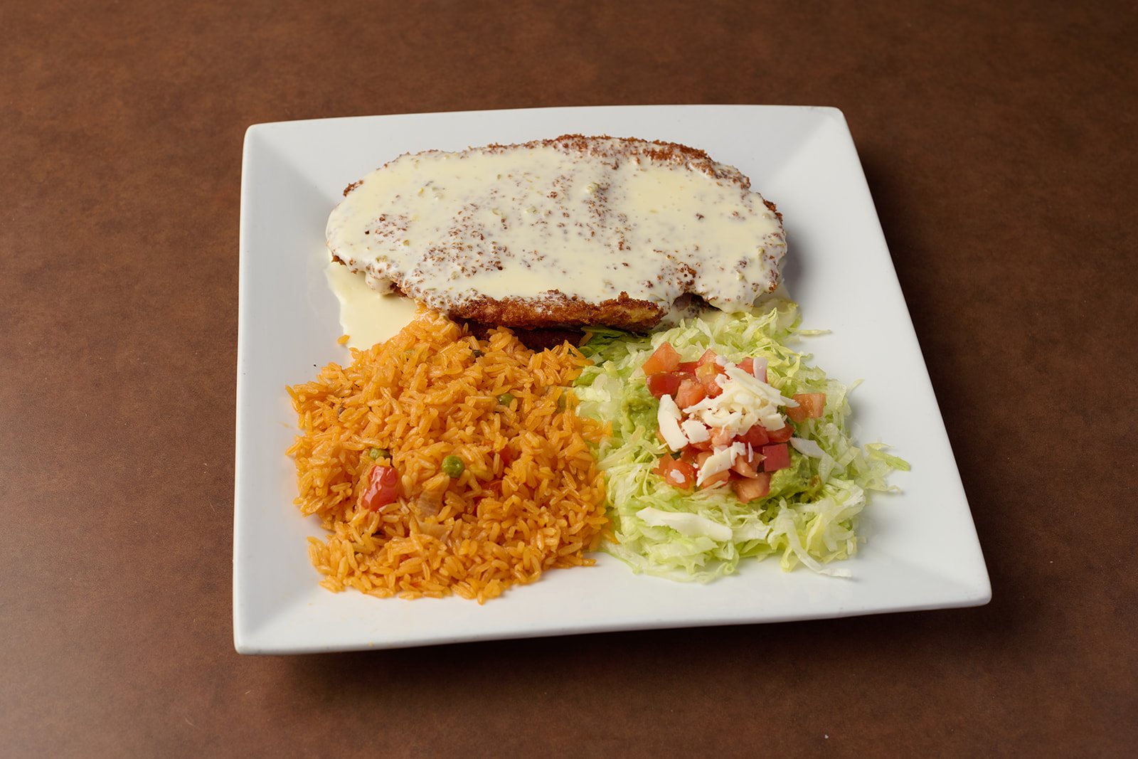 Plate with breaded fried fish covered in white creamy sauce, serving of seasoned rice with peas and diced tomato, and shredded lettuce salad with chopped tomatoes and shredded cheese.