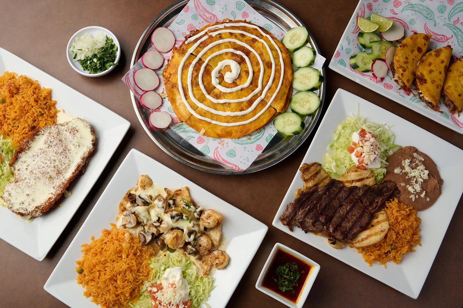 A variety of Mexican dishes on white plates, including a chicken fried steak with white sauce, a plate of shrimp with mushrooms and rice, grilled steak with sides, cheese quesadillas, and an after-meal flan with cucumber slices, radishes, and lime we