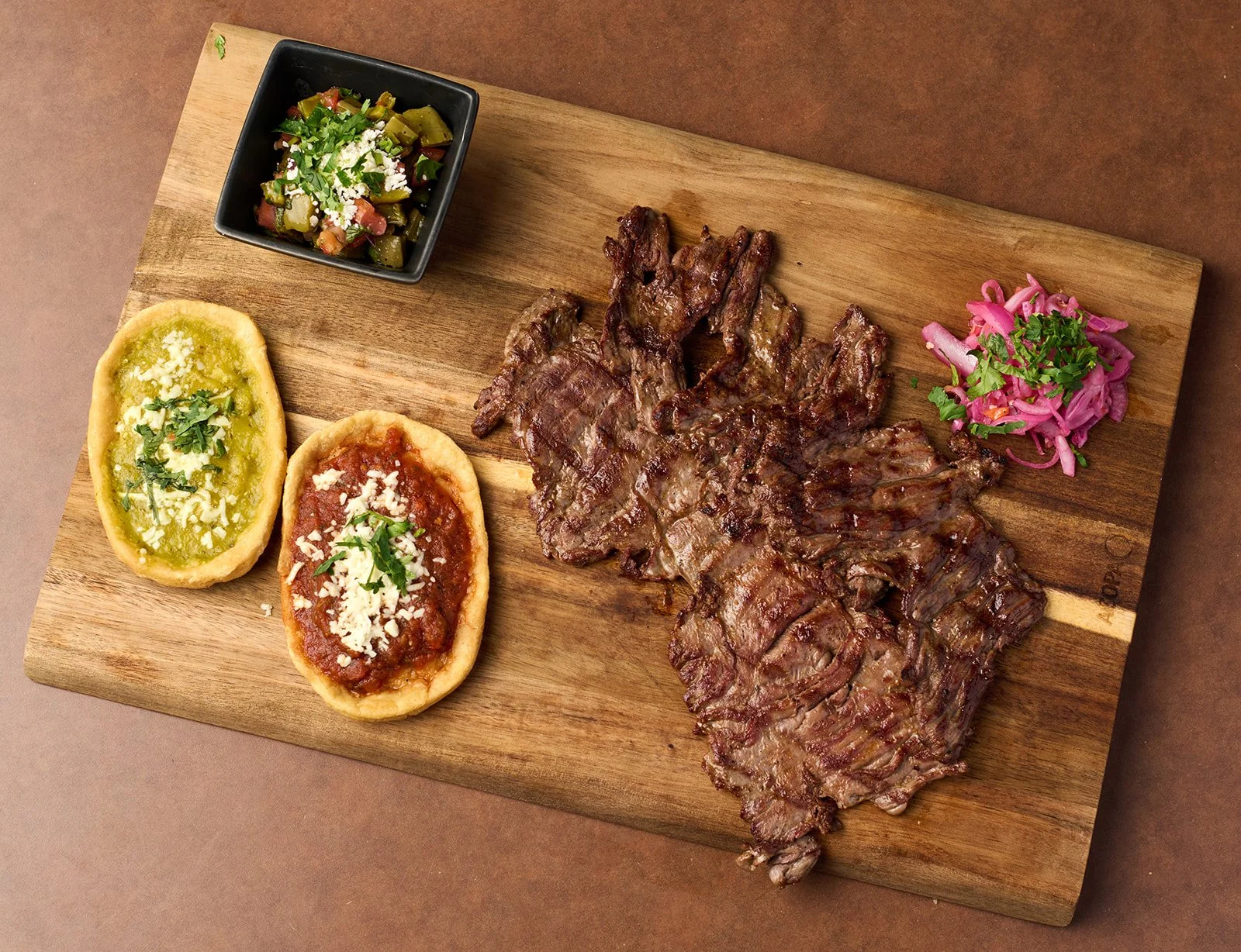 Grilled beef steak with pink pickled onions, two mini tacos with cheese and sauce, and a side of chopped vegetables in a small black dish on a wooden cutting board.
