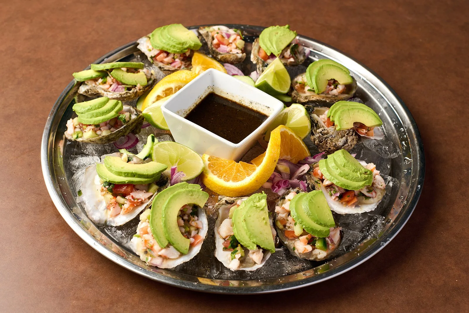 A round metal tray of half-shell oysters topped with chopped vegetables, slices of avocado, and lime wedges, with a small container of dark dipping sauce in the center.