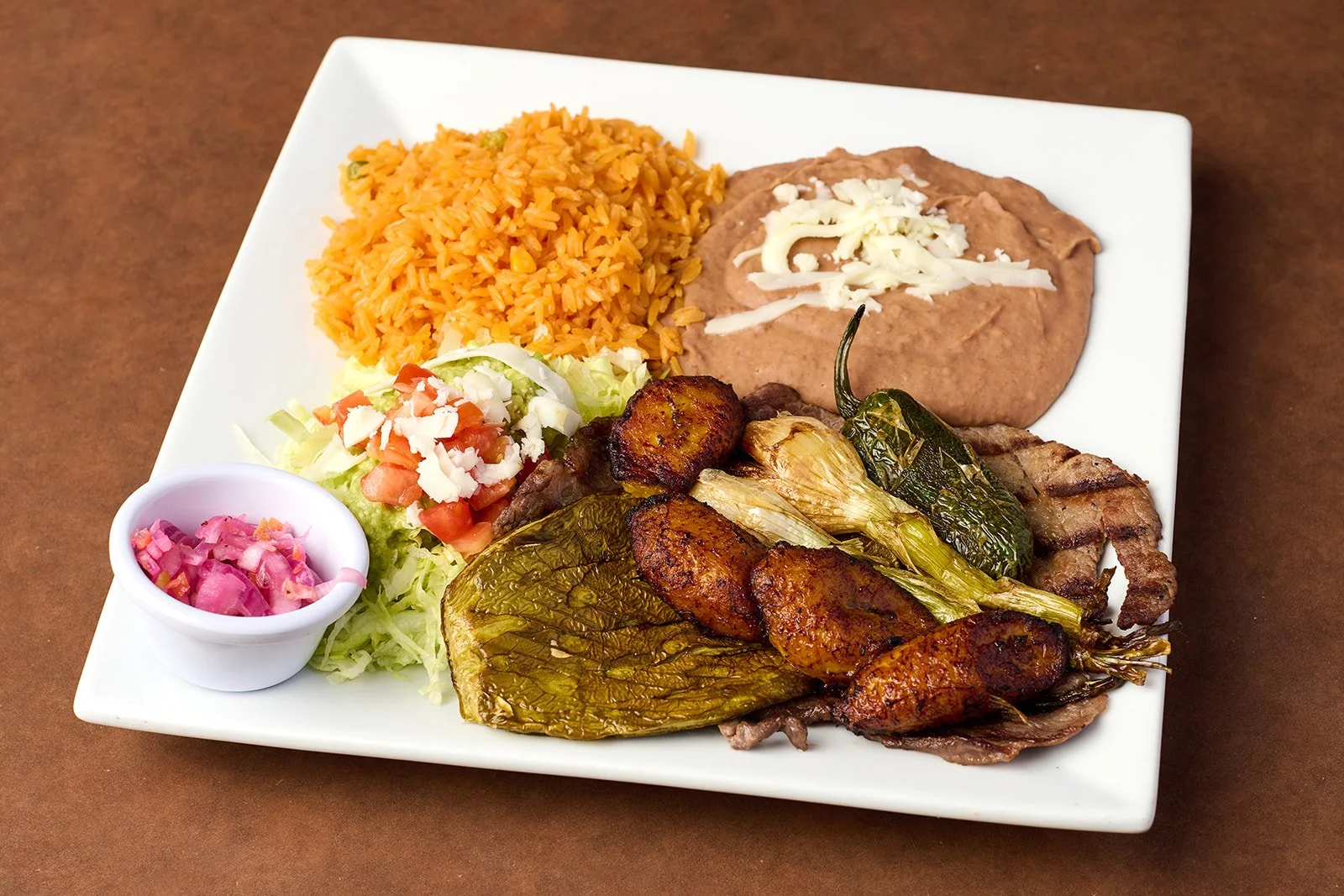 Plate of Mexican food with rice, refried beans with cheese, grilled meats, roasted poblano pepper, fresh salad, and pink pickled onions.