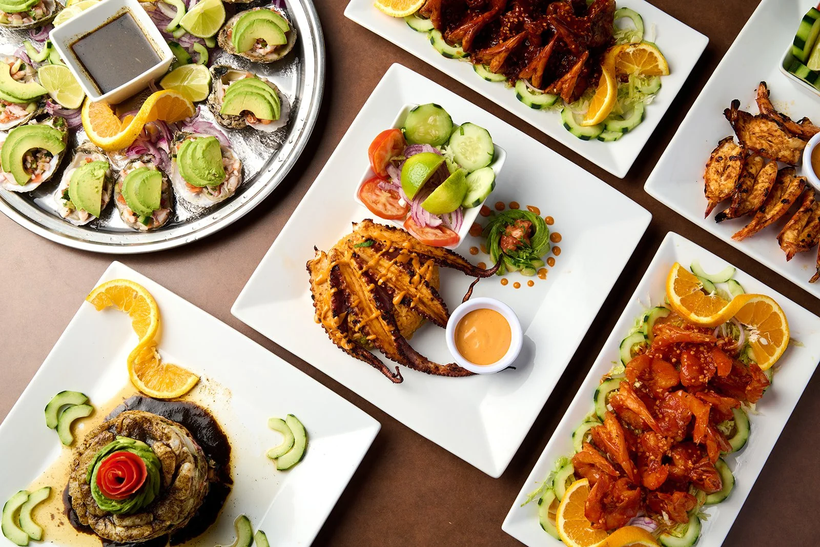Assorted Asian and Japanese dishes on white and silver platters, including sushi with avocado and lemon slices, grilled chicken wings, spicy fish or chicken salad, and grilled octopus with vegetables, garnished with vegetables, orange slices, and sau