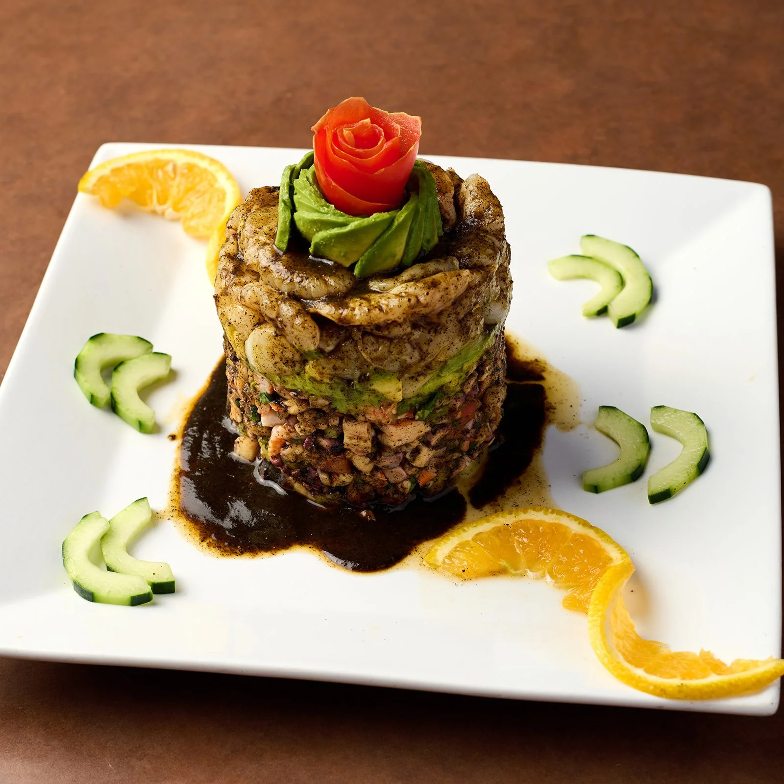 Layered sushi dish on white plate with cucumber slices, orange wedges, and soy sauce.