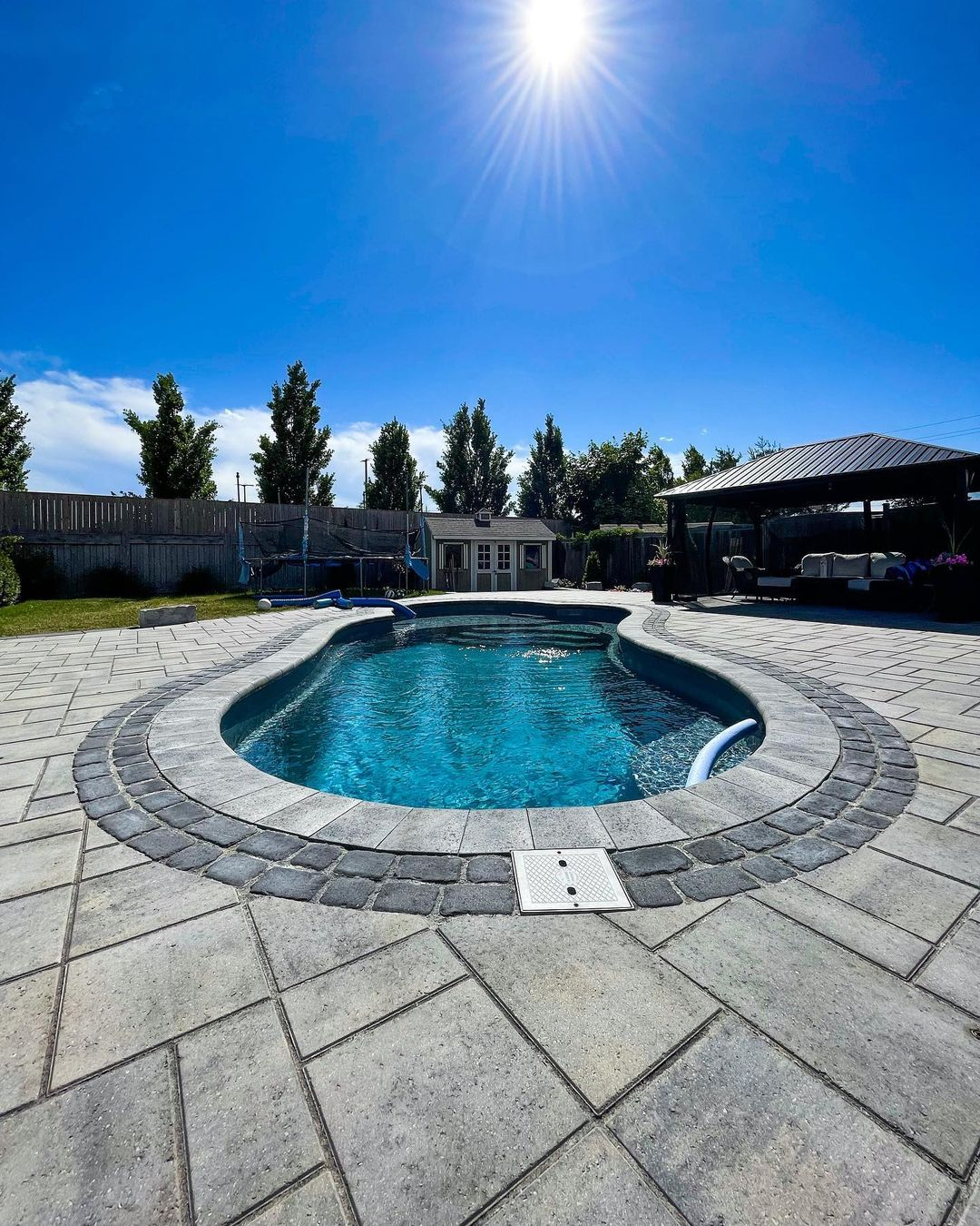 A backyard with a swimming pool under a bright, sunny sky. There is a fenced yard with trees, a trampoline, a shed, and a shaded seating area with outdoor furniture.