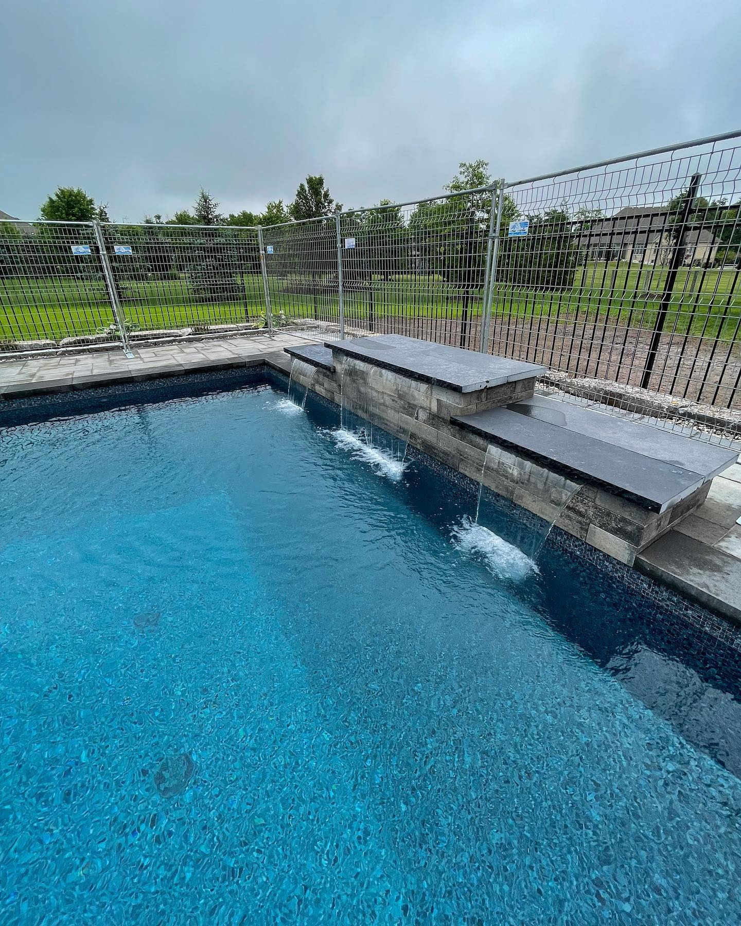 A backyard swimming pool with a modern stone waterfall feature, surrounded by a metal fence and a green lawn with trees in the background.