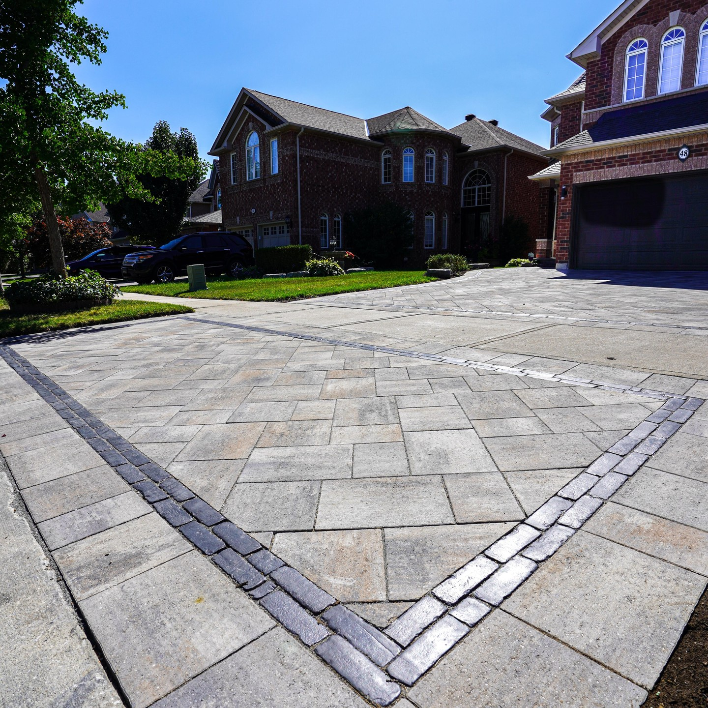 A residential driveway with patterned concrete pavers, leading to a large brick house with multiple windows and a garage, surrounded by green grass and trees.
