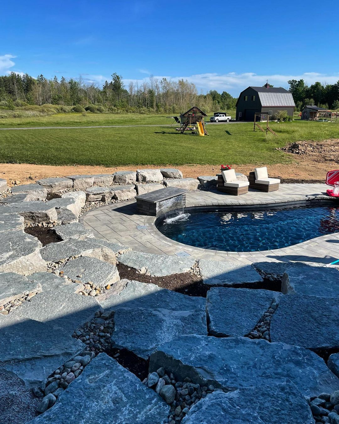 View of a backyard with a swimming pool surrounded by stone pavers and chairs, a grassy lawn with outdoor play equipment, and a distant barn and trees under a clear blue sky.