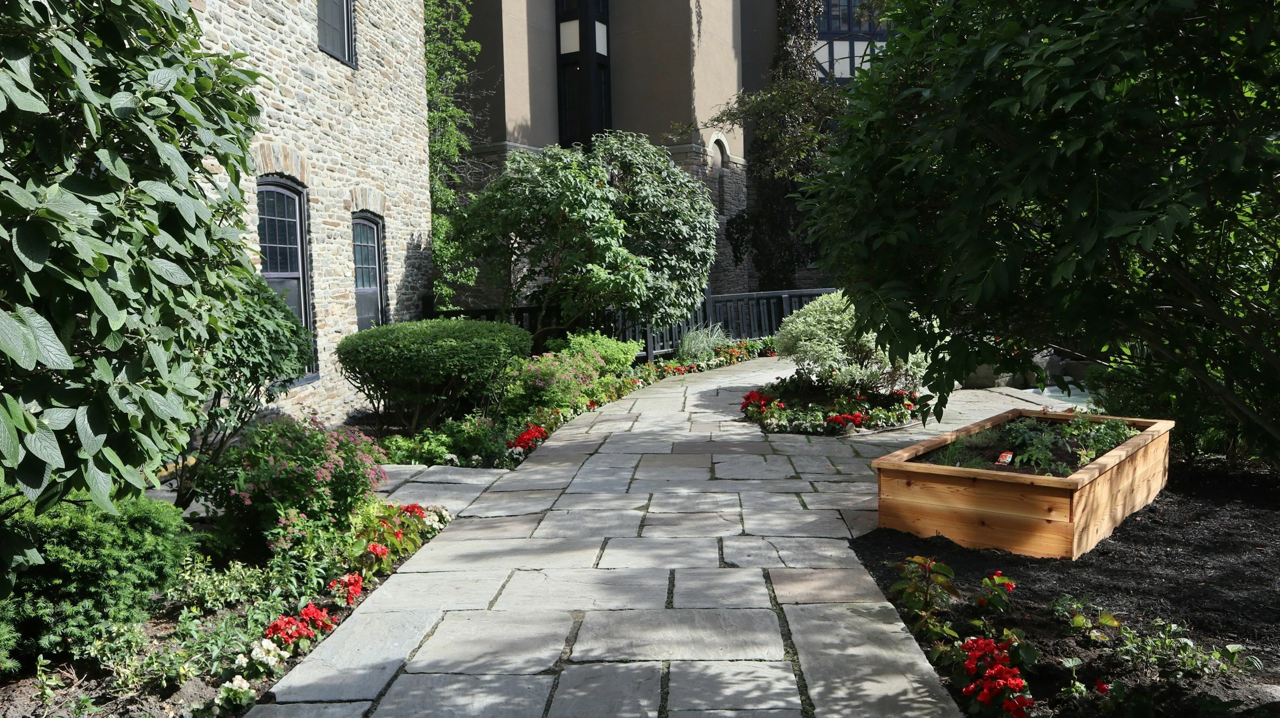 A stone pathway winds through a lush garden with various green bushes, flowering plants, and trees, next to a stone building with black window frames and a beige wall.