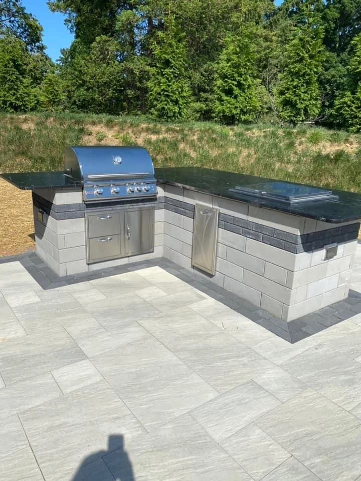 Outdoor kitchen with a grill, a covered cooking area, and a flat surface for preparing food, built with light-colored brick, black accents, and stainless steel appliances, set in a paved area with green trees in the background.