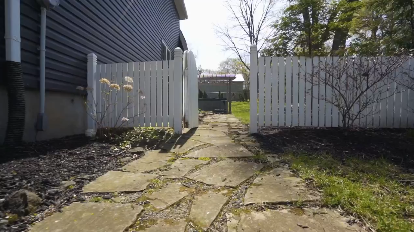 View of a backyard pathway made of irregular stone slabs, leading through a white picket gate to a grassy area with trees, a shed, and a house in the background.