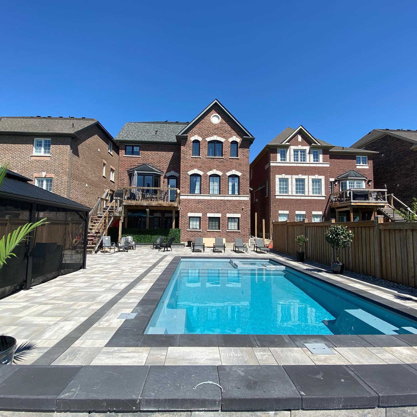 A backyard with a swimming pool surrounded by lounge chairs, potted plants, and a fenced area. There are three multi-story brick houses in the background under a clear blue sky.