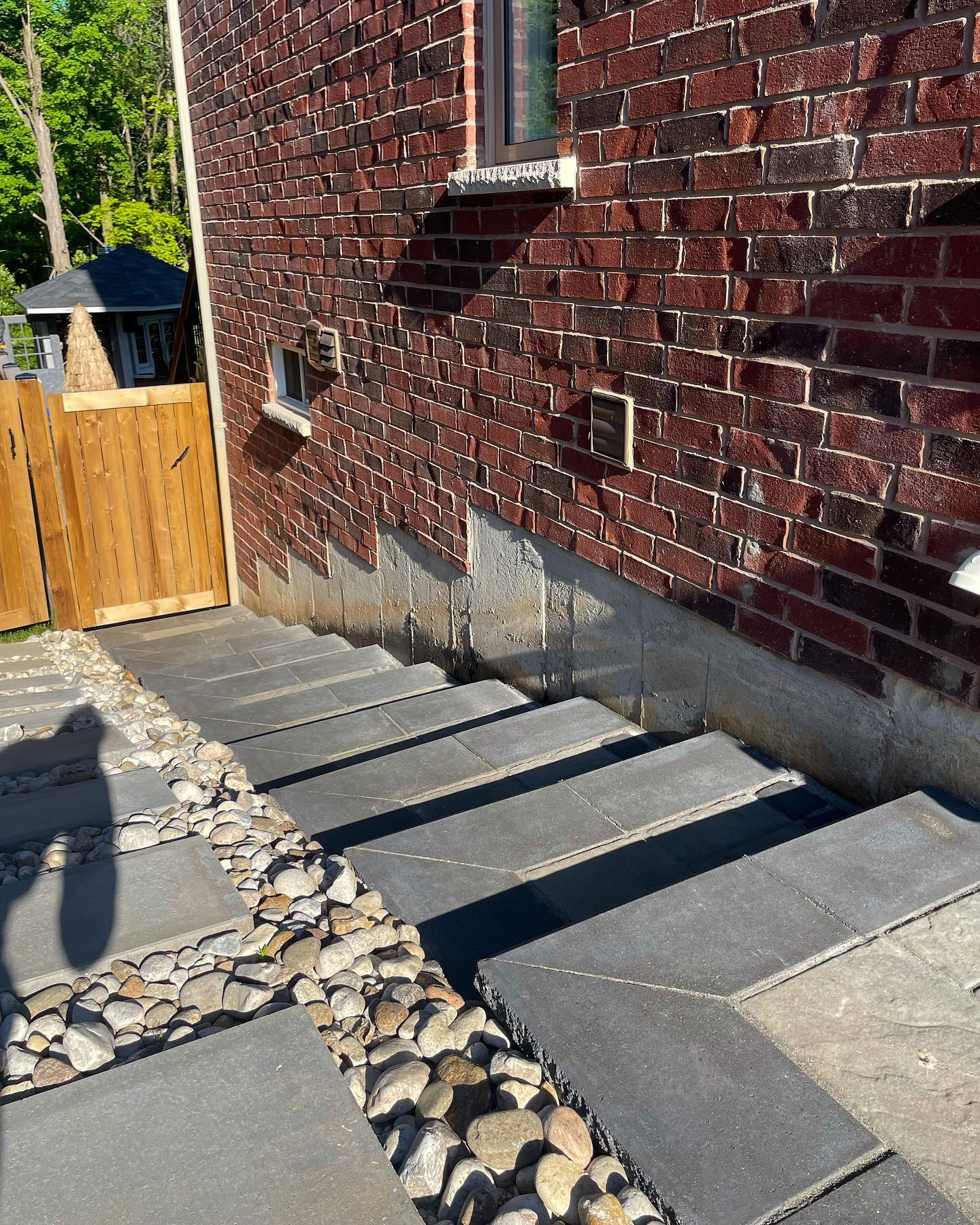 Backyard stairs leading up to a brick house with two small windows, a wooden fence gates on the left, and a pebble-lined area in front of the stairs.