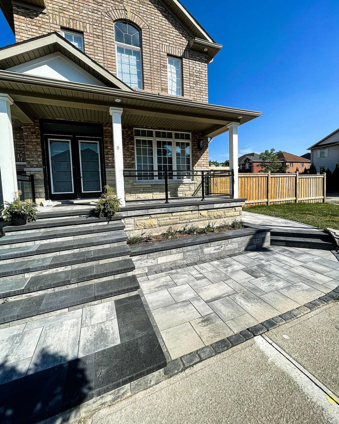 This is a photo of the front of a two-story brick house with a stone staircase leading to a small porch. The porch has black railings and two potted plants. There are two doors on the porch, and a large window with multiple panes. The house has a sloped roof, and there is a fenced yard with a small lawn. The sky is clear and blue.